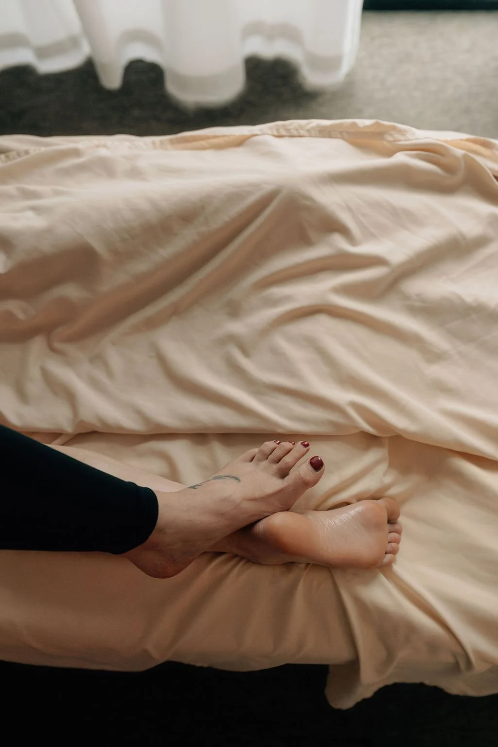 Person with painted toenails sitting on a bed with beige sheets, showing one foot on top of the other.