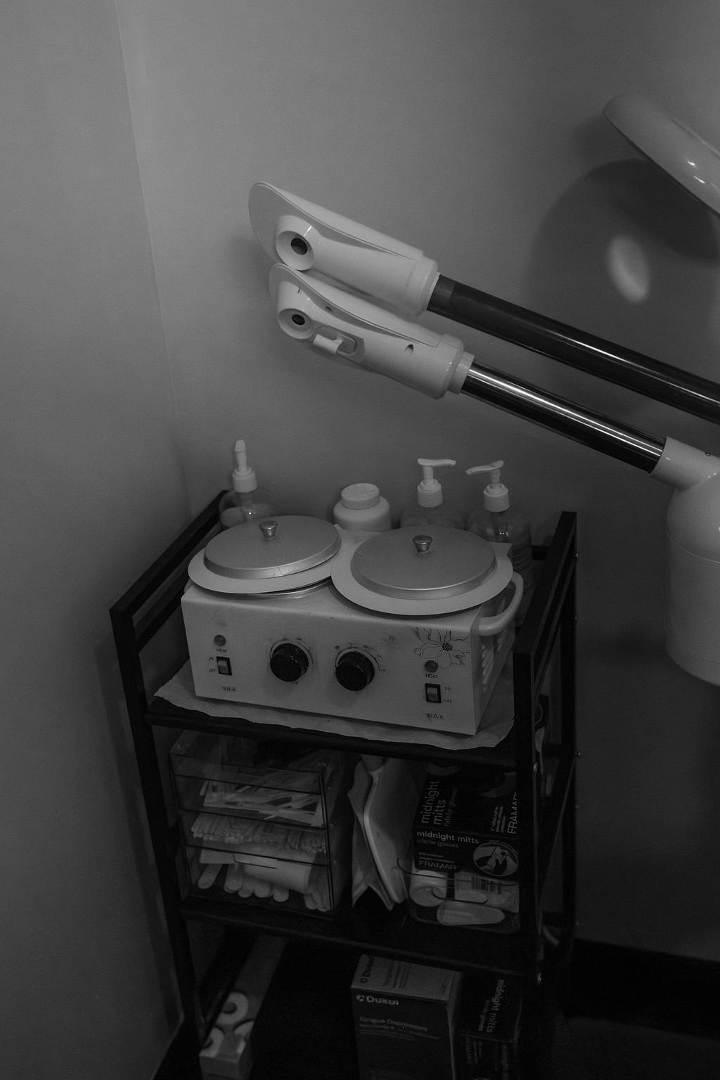 A black and white photo of a small spa or facial treatment room. There are two large robotic facial steamers mounted on the wall above a small black metal cart. The cart holds various skin care products, steamers, and supplies, including bottles and boxes. The room has minimal furniture and appears to be a specialized treatment area.