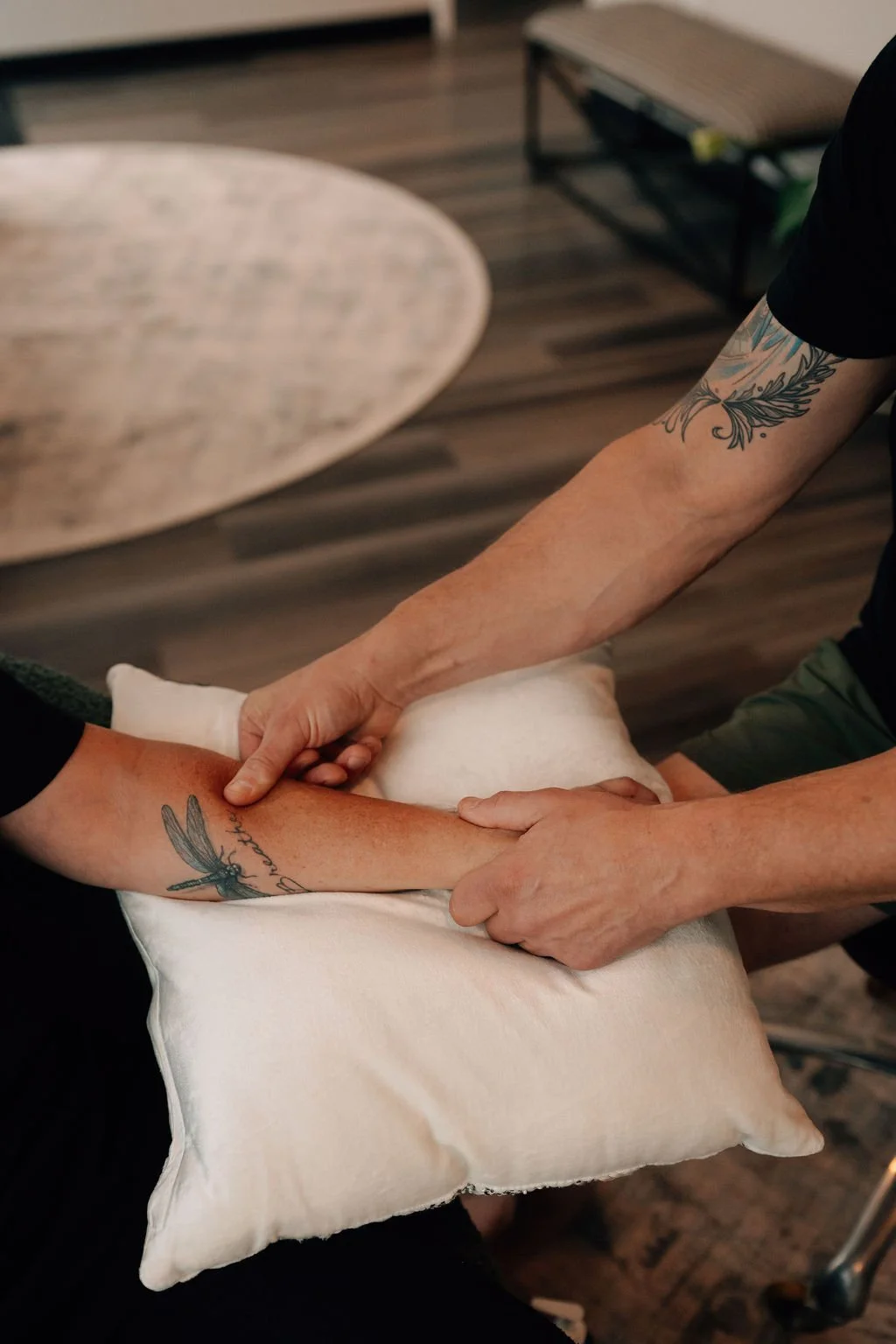 Two people holding hands over a pillow, with tattoos on their forearms, in a room with wooden flooring and a round table.