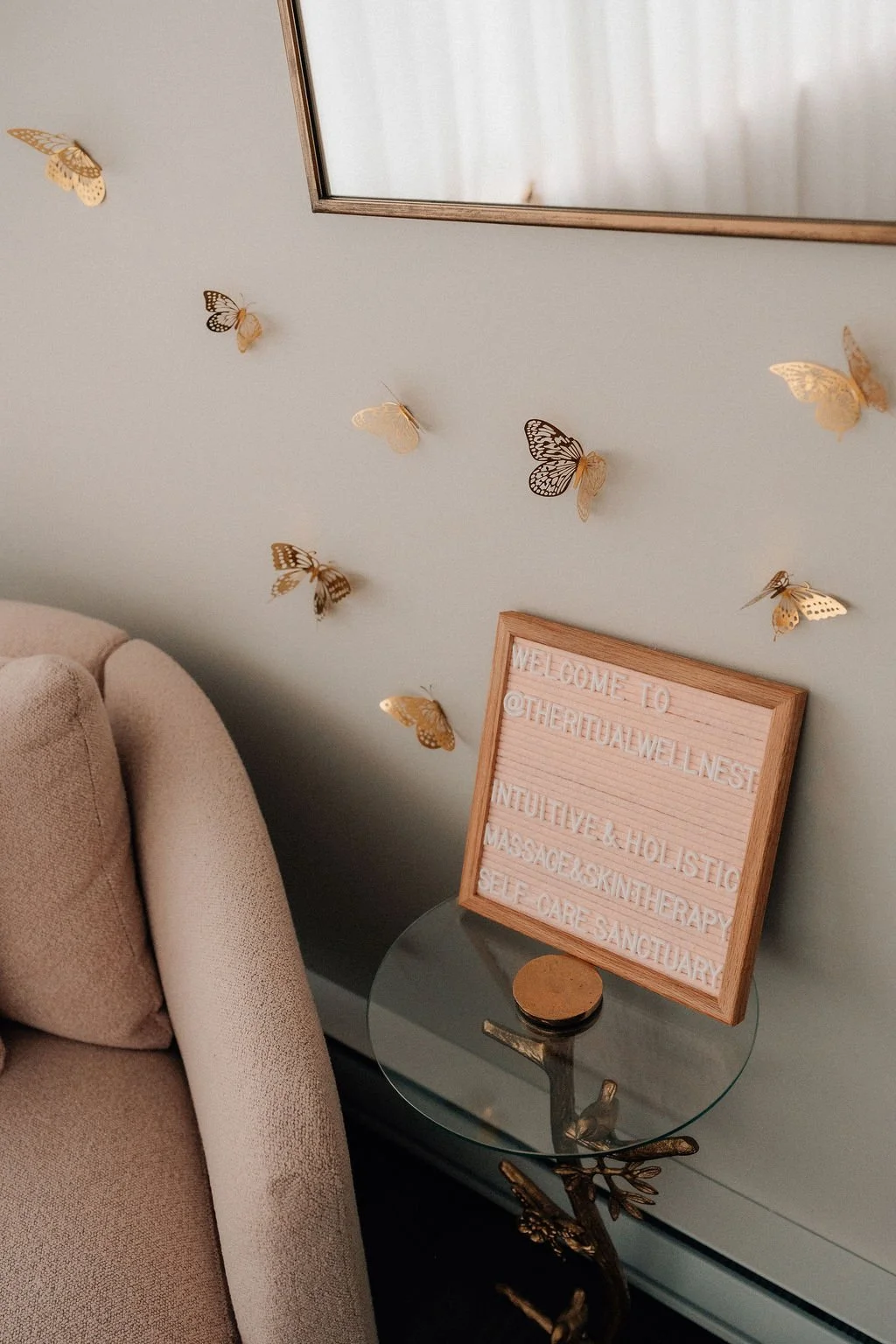 Decorative wall with gold butterfly decals, a framed sign with social media handle and wellness services, a glass side table with a decorative gold element, and part of a beige sofa.