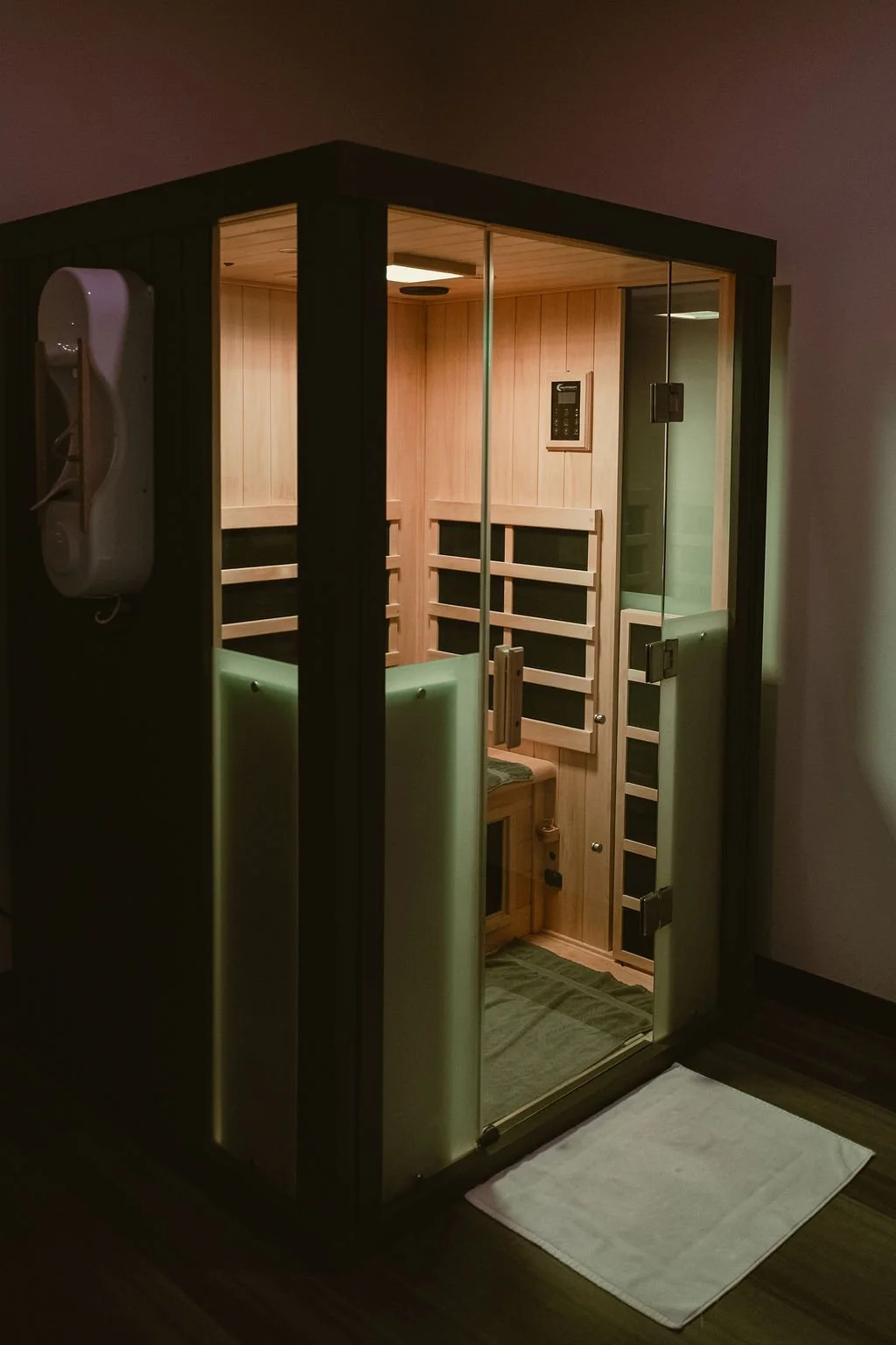 A modern infrared sauna with glass doors and wooden interior, featuring a small bench, control panel, and a telephone mounted outside the front.