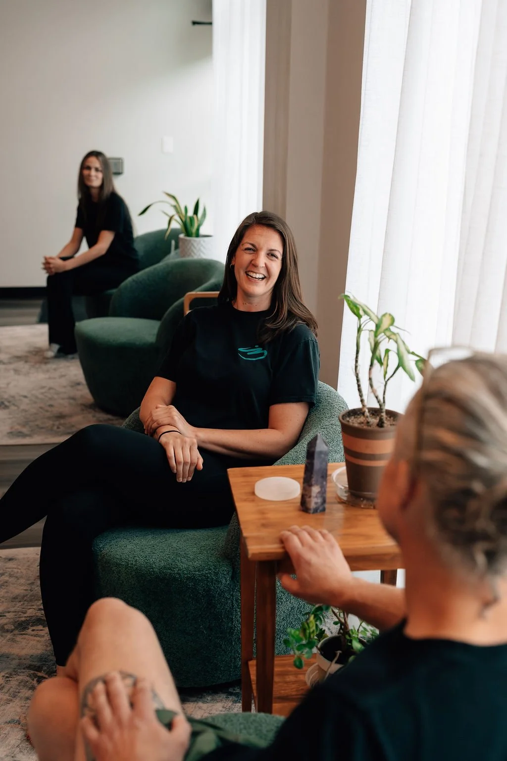 Three women in a cozy lounge area with green armchairs, potted plants, and natural light. One woman with brown hair smiling directly at the camera, another woman with brunette hair in the background, and a third woman with gray hair partially visible in the foreground.