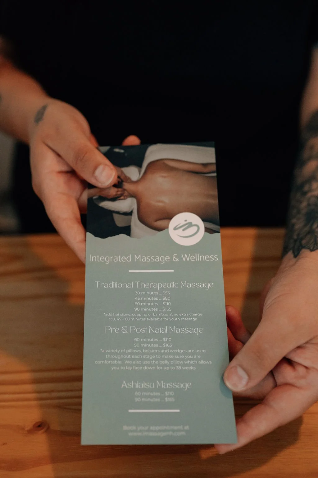Person holding a massage service brochure on a wooden table, partially visible tattooed arms, black clothing, and a tattoo on the left wrist.