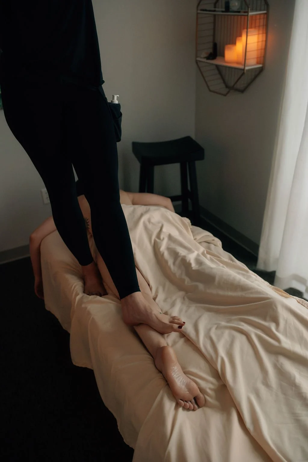 A person is lying on a bed covered with beige sheets, with legs and feet visible. Another person, dressed in black with a hand sanitizer attached to their pocket, is standing on the bed, stepping over the person lying down. The room has a dark wooden floor, a small black chair, a wall-mounted shelf with candles, and a window with white curtains.