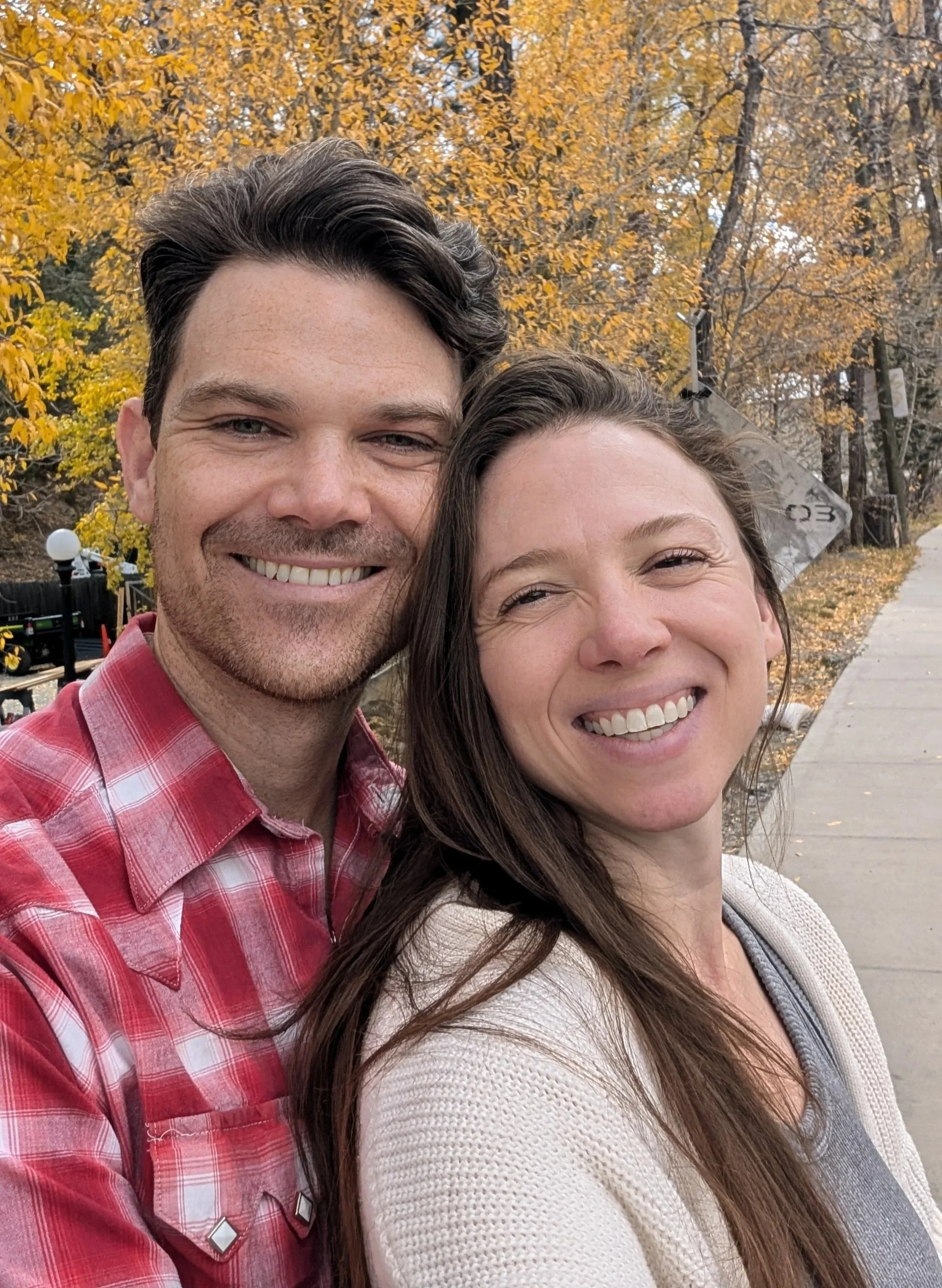 A smiling couple taking a selfie outdoors during fall with colorful yellow and orange trees in the background.