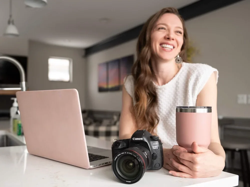 Smiling woman sitting at a kitchen counter with a pink laptop, a Canon camera, and a pink tumbler.