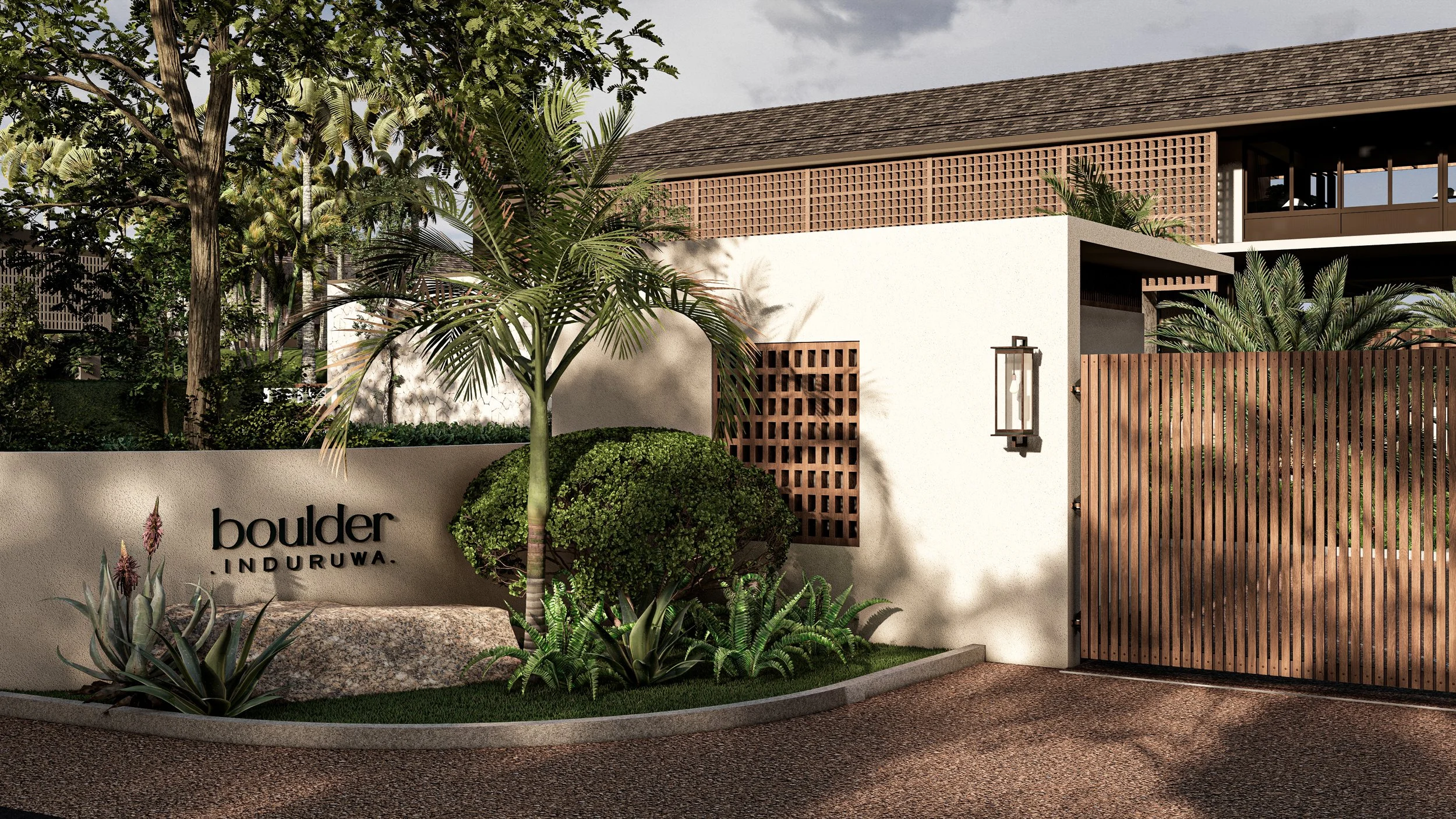 Entrance gate with wooden fencing and lush tropical plants, sign reads 'boulder Induruwa' in front of a modern building with a sloped roof and greenery.