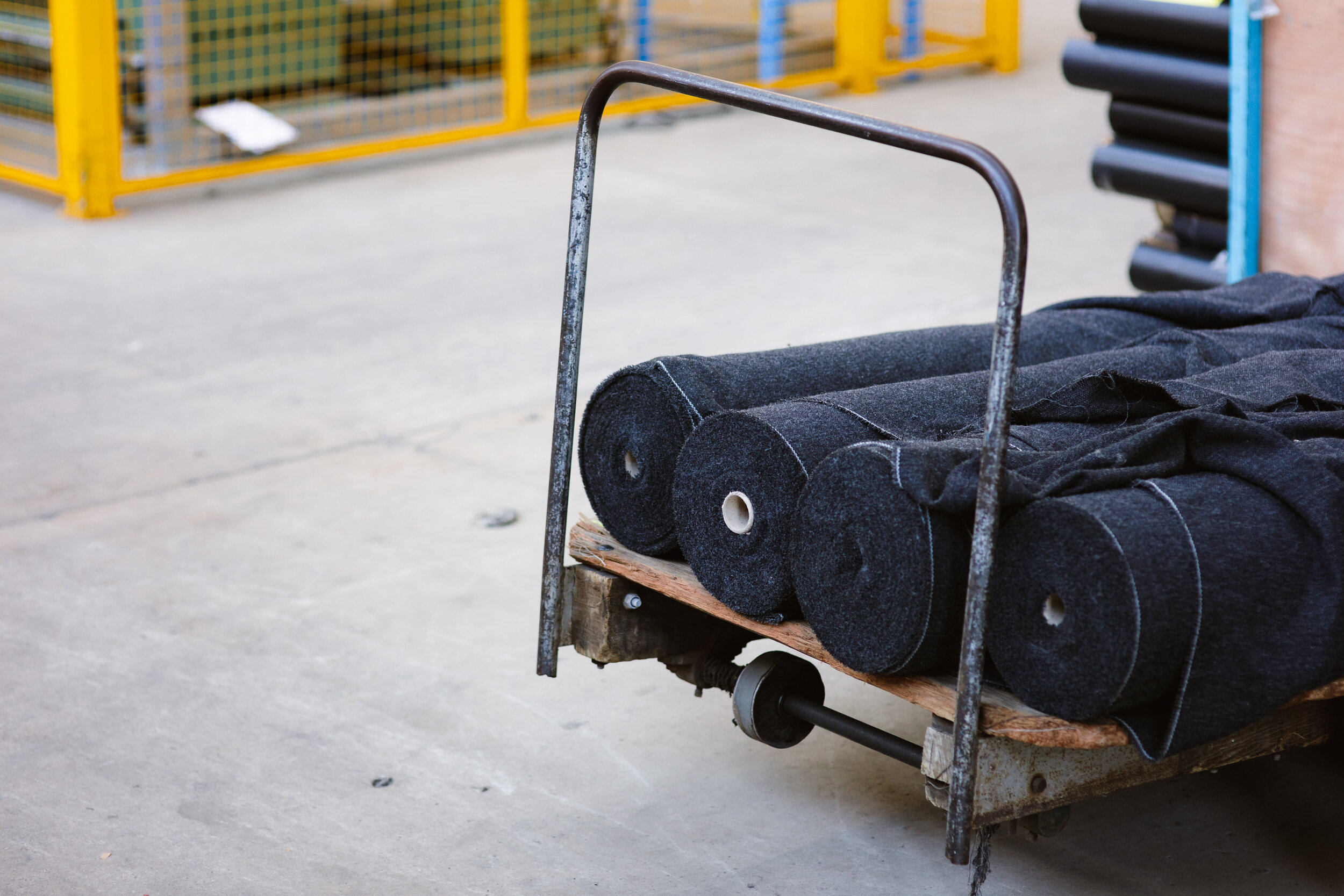 A metal cart with black fabric rolls stored on a wooden pallet, located in a warehouse or industrial setting, with yellow safety barriers in the background.