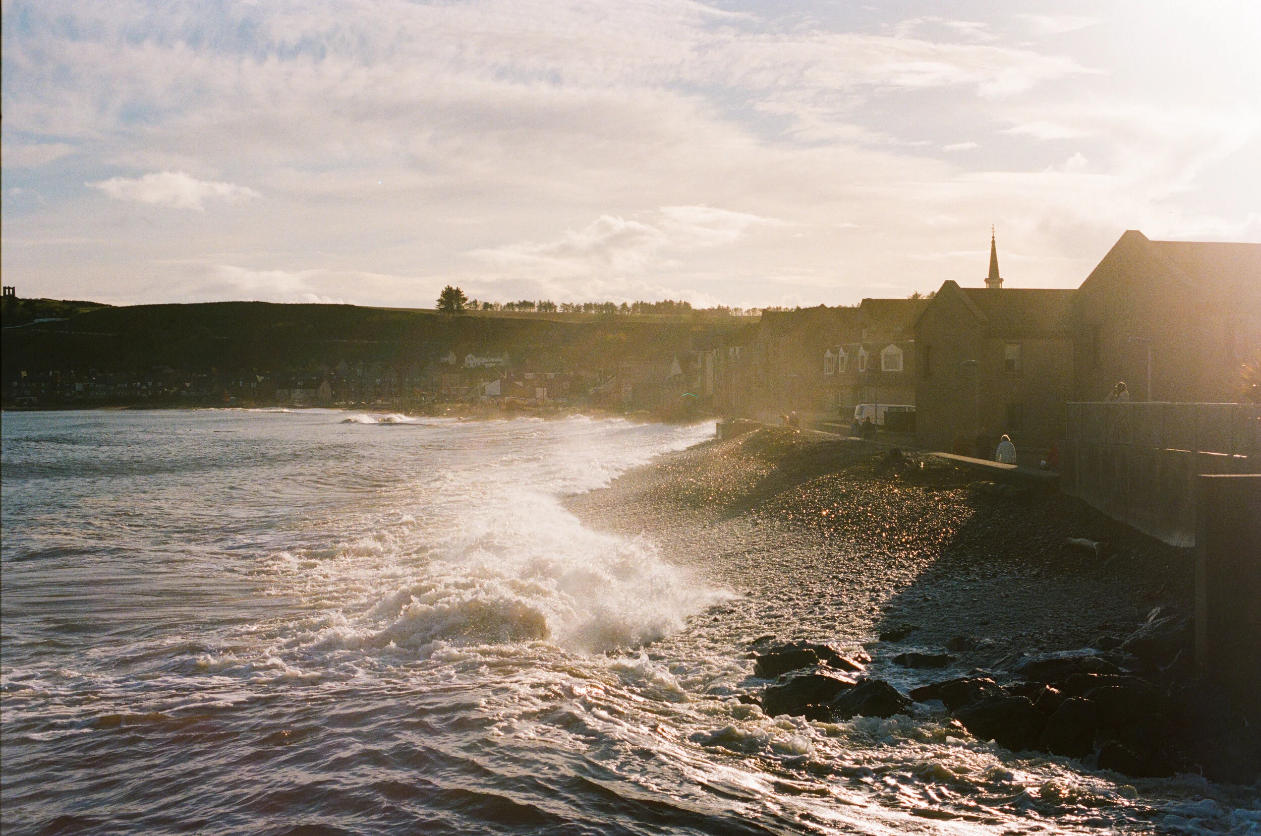A scenic view of a coastal village with houses, a church steeple, and grassy hills under a partly cloudy sky, taken at sunset or sunrise with sunlight reflecting on the water.