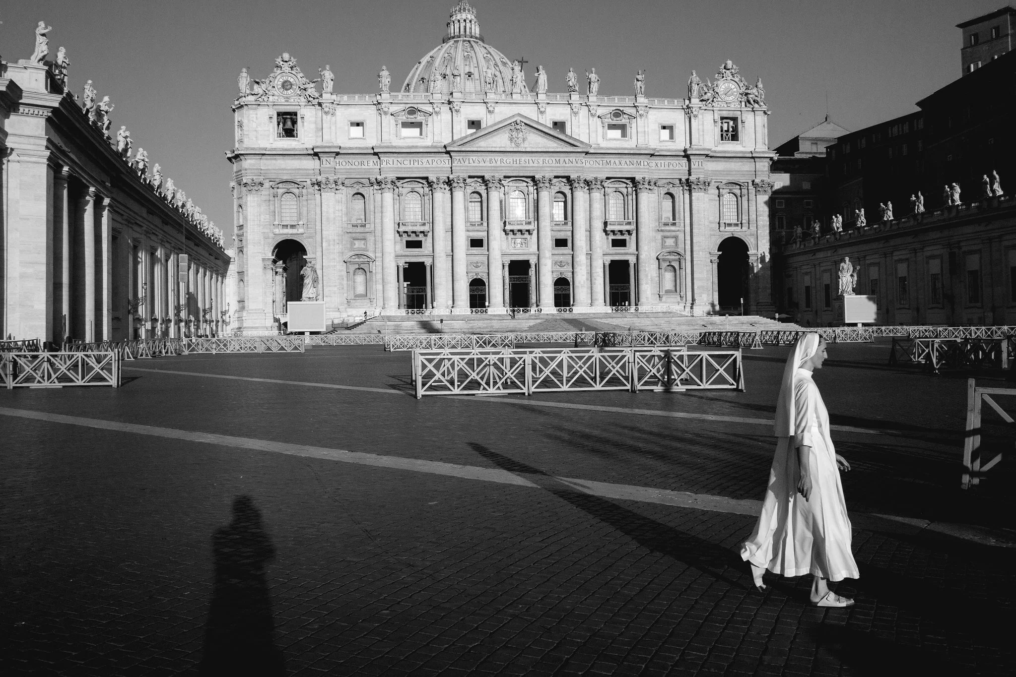 A black-and-white photograph of St. Peter's Basilica in Vatican City with a nun in religious attire walking across the square, and long shadows cast on the cobblestone ground.