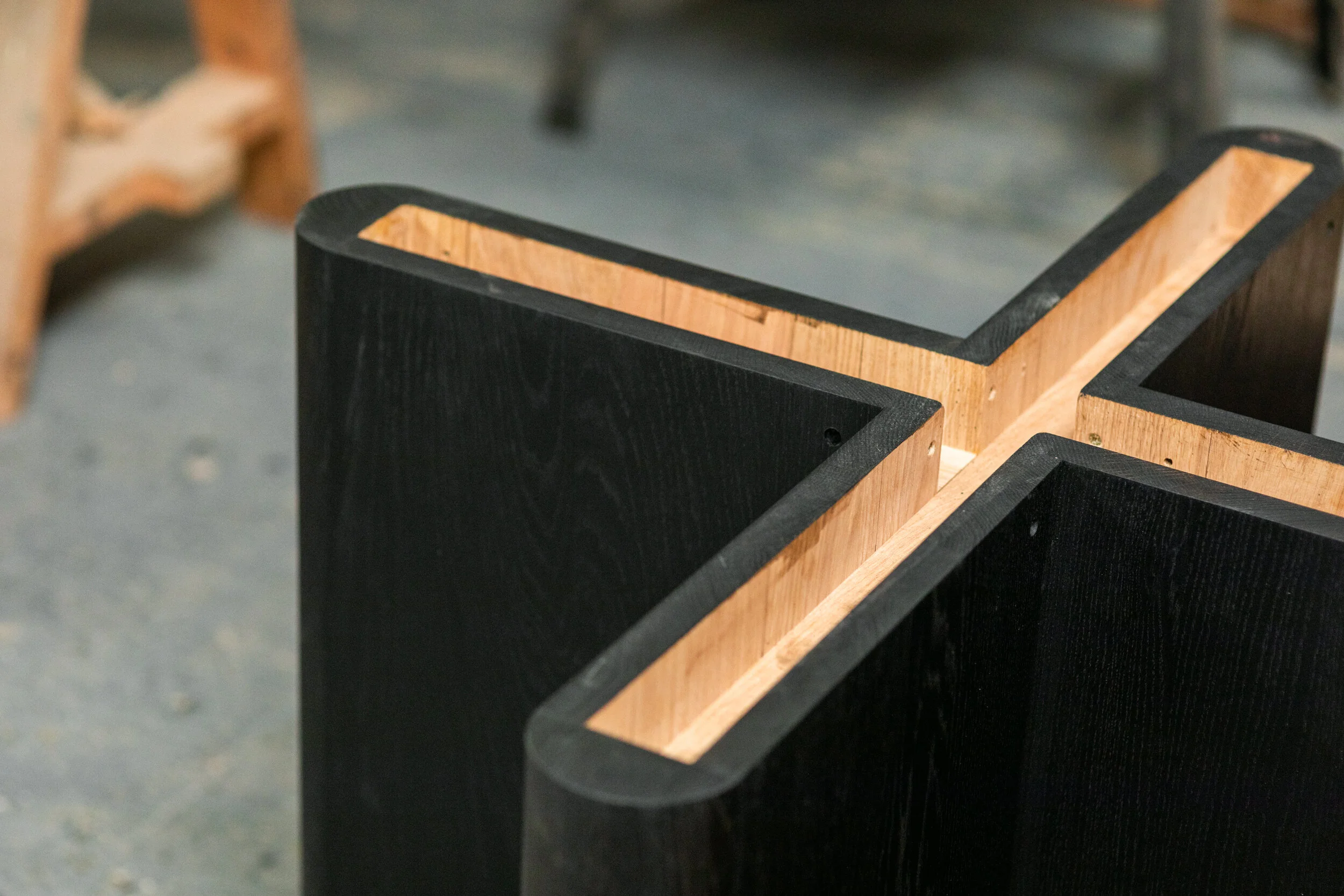 Close-up of wooden and black-painted structural pieces, possibly furniture or construction components, placed on a concrete floor, with some blurred wooden furniture in the background.