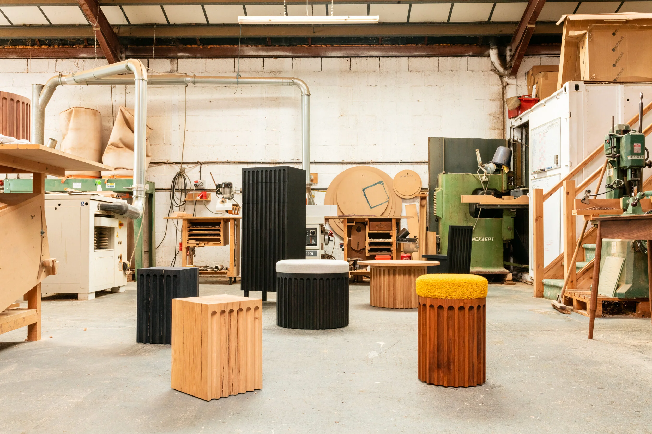 Workshop or studio space with wooden furniture and tools. Various wooden stools, cabinets, and cutouts are scattered around. The industrial setting features a white brick wall and visible metal beams ceiling.