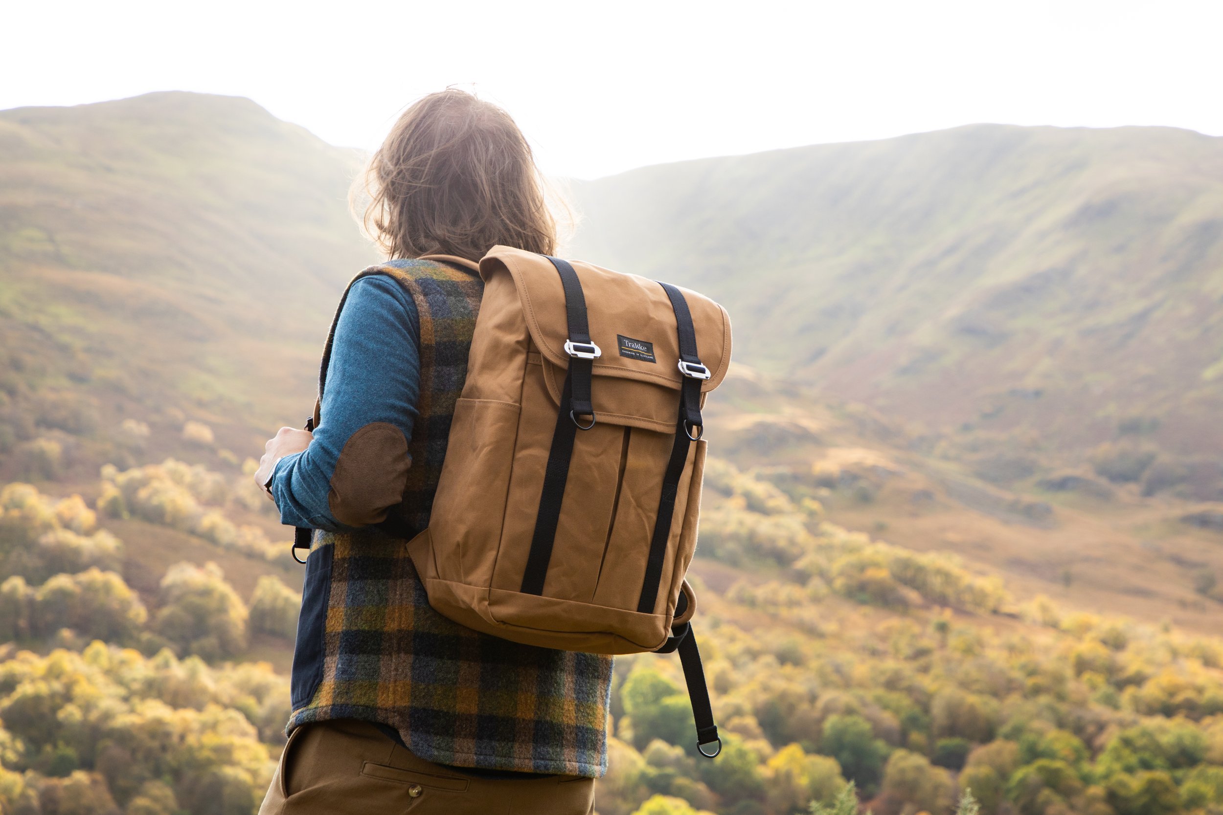 Person with shoulder-length hair wearing a plaid jacket and brown pants, carrying a tan backpack, standing outdoors overlooking rolling green hills and a bright sky.