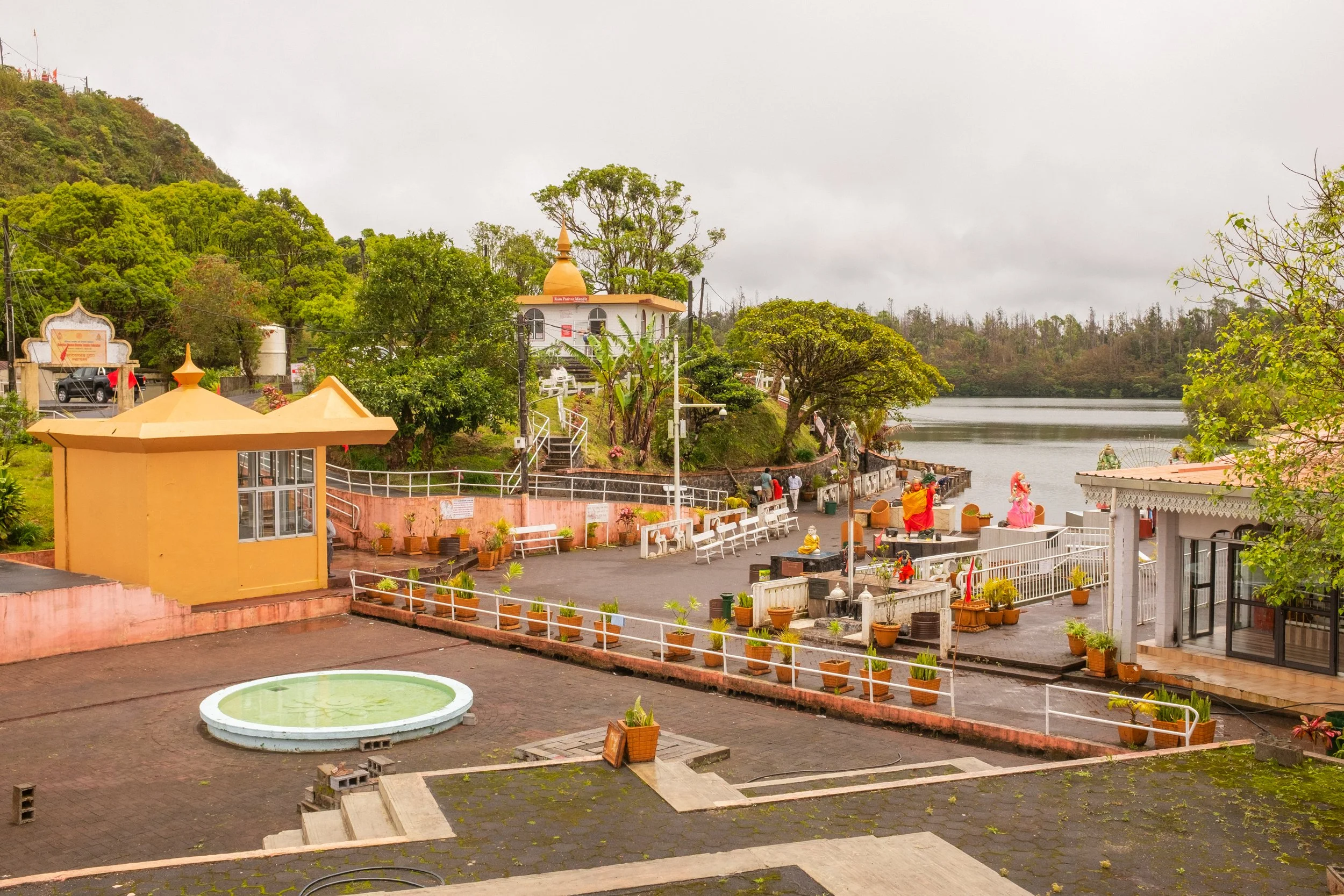 A scenic view of a temple complex situated beside a river, surrounded by green trees and hills. There are several buildings with orange and white architectural elements, small statues, potted plants, and sitting benches near the water. The sky is ove