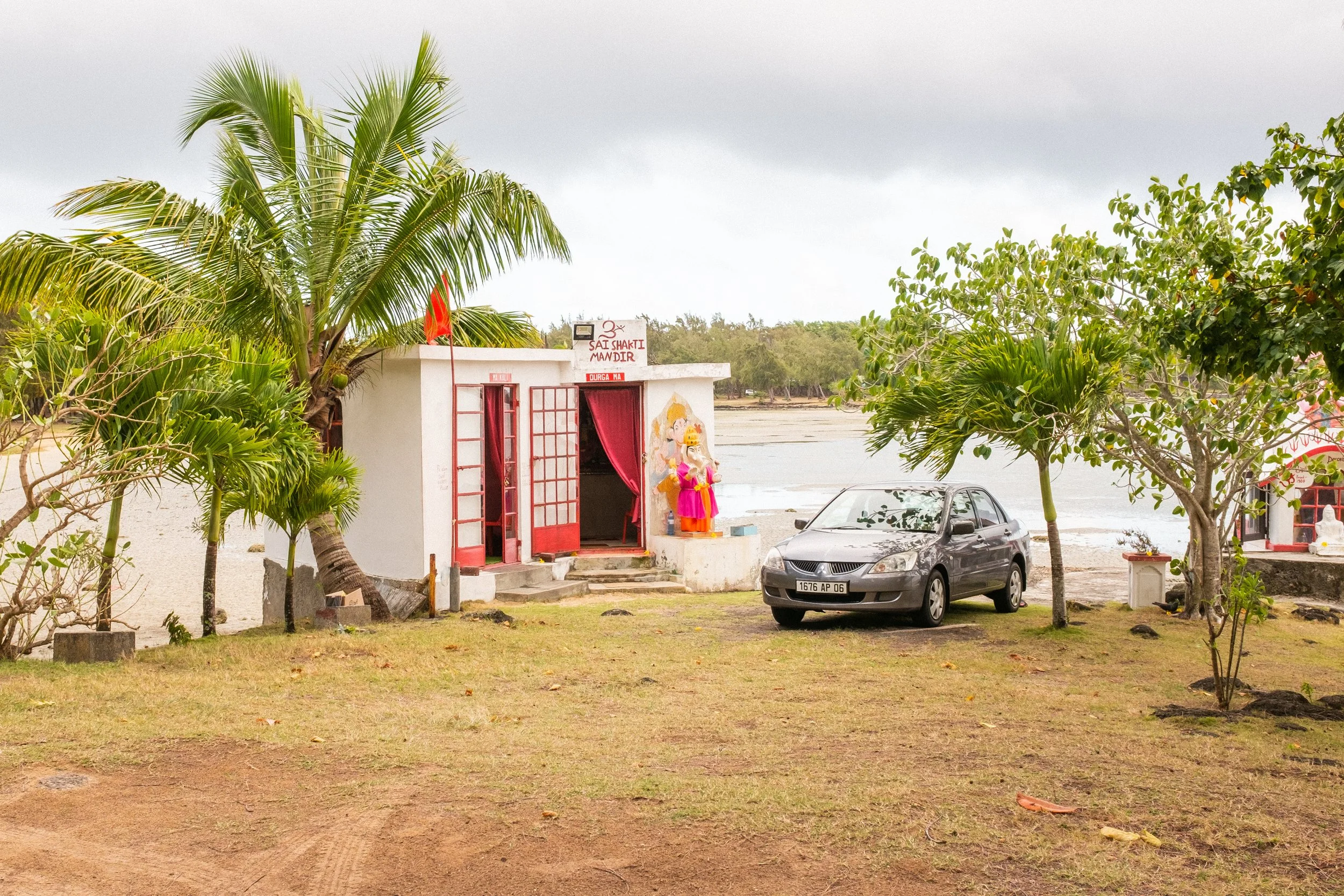 A small building with Hindu deity statues and a sign that reads 'Sai Shakti Mandir' at the beach, surrounded by greenery, a car parked nearby, overcast sky in Mauritius