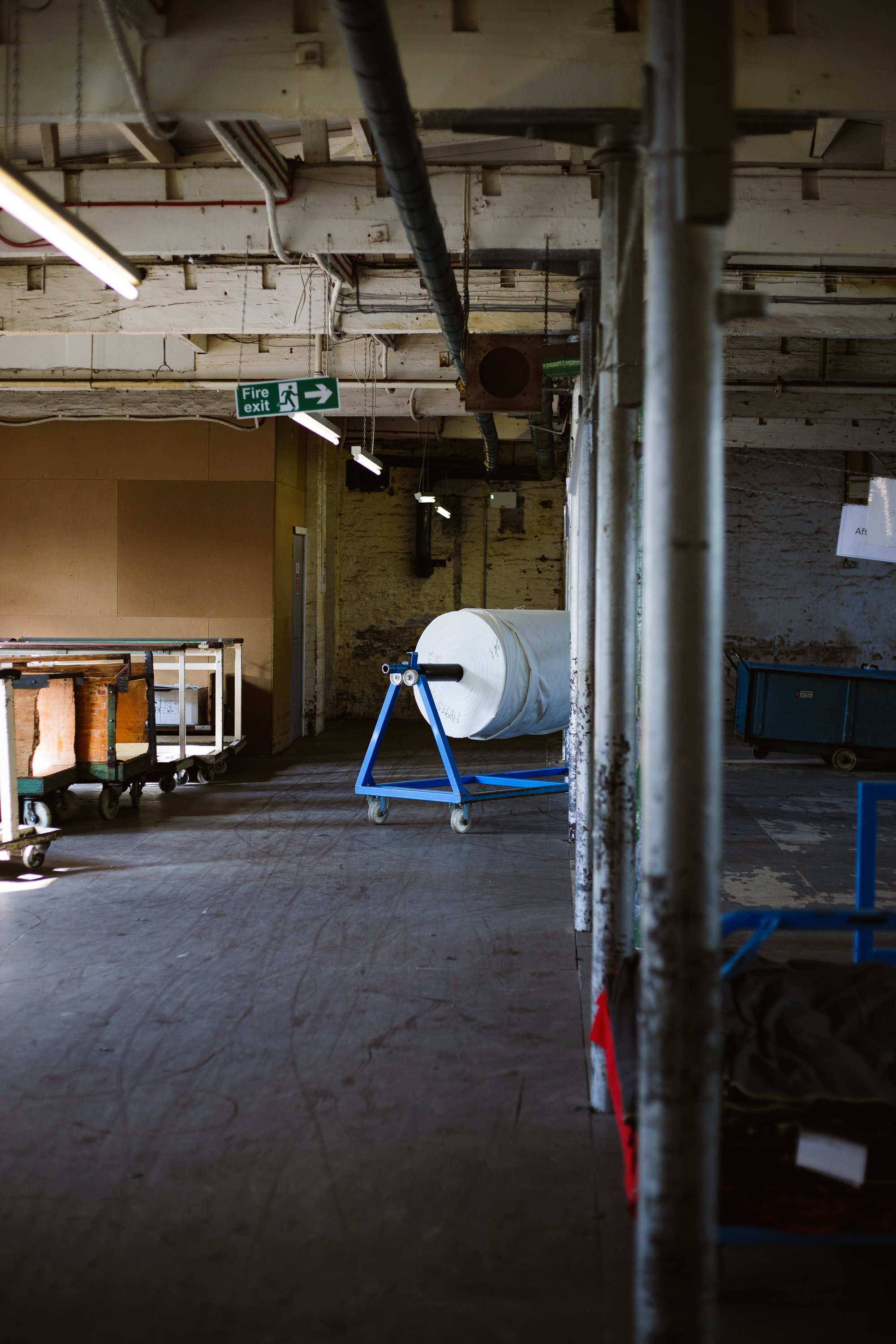 Industrial space with scaffolding, pipe vents, and a large roll of paper on a blue stand, illuminated by overhead lights. A green fire exit sign points to the right.