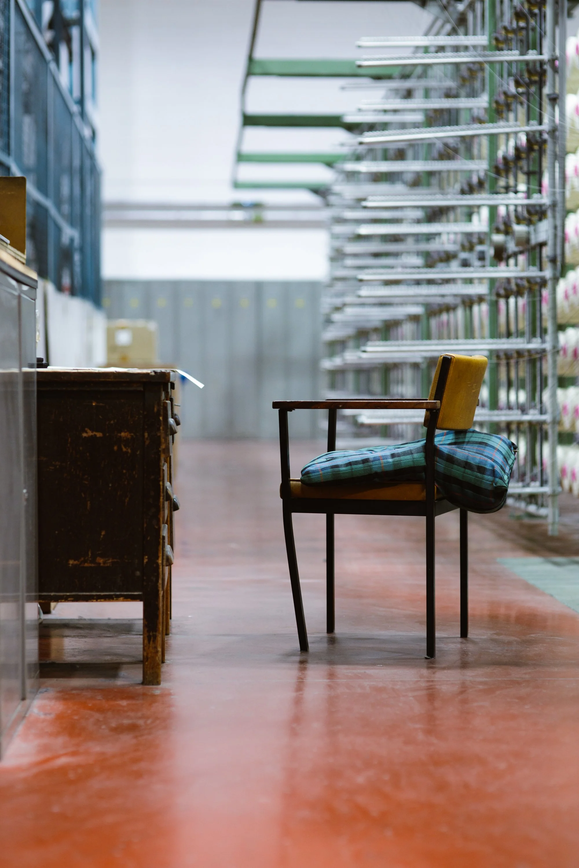 Empty store aisle with a yellow chair, stack racks to the right, and a worn brown cabinet on the left, with a polished reddish-brown floor.