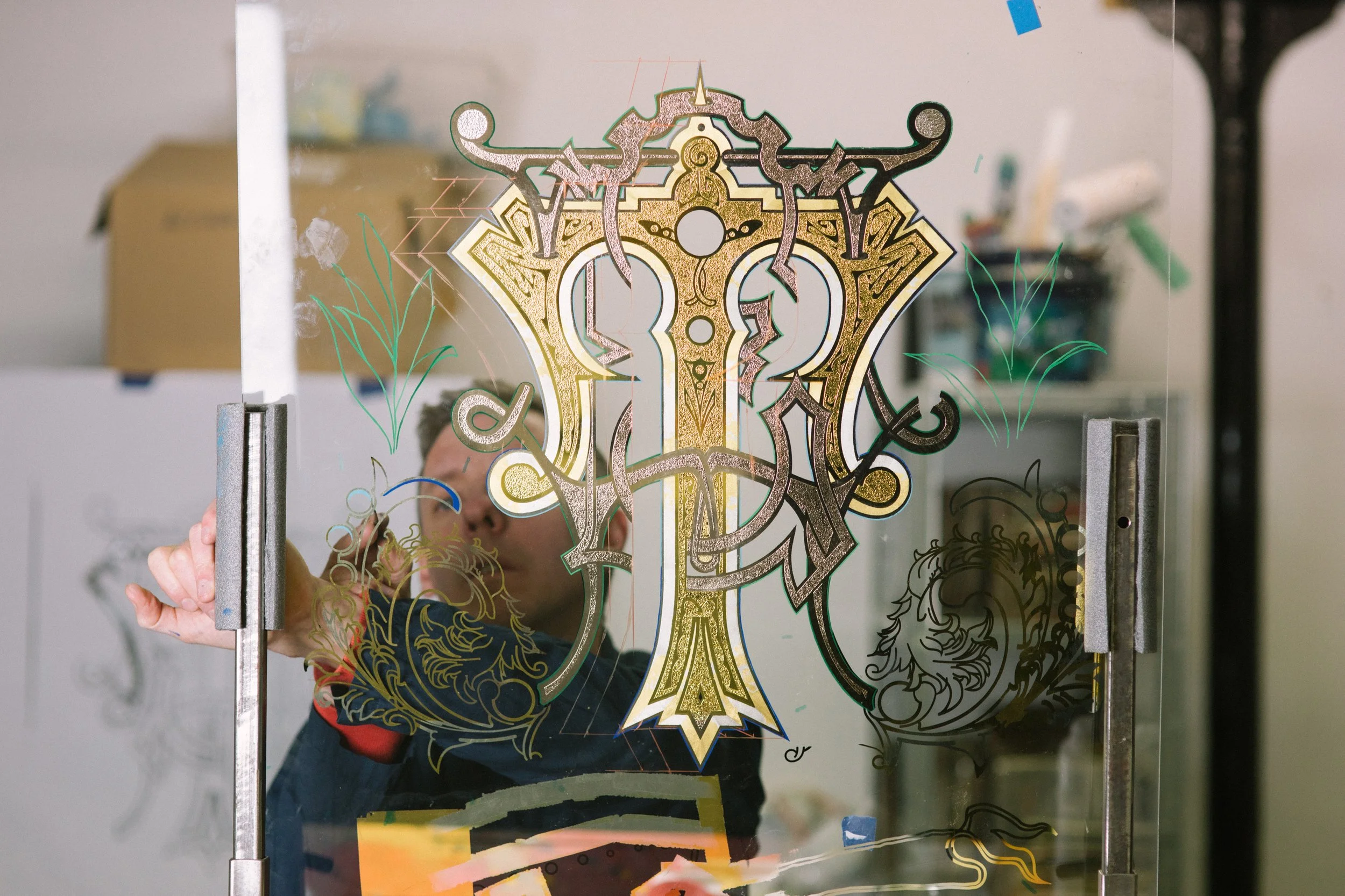 A person is installing or working on a decorative glass panel featuring an intricate gold and black design of a stylized figure with symmetrical patterns, floral motifs, and lightning-like elements. The person is partially visible, holding the panel 