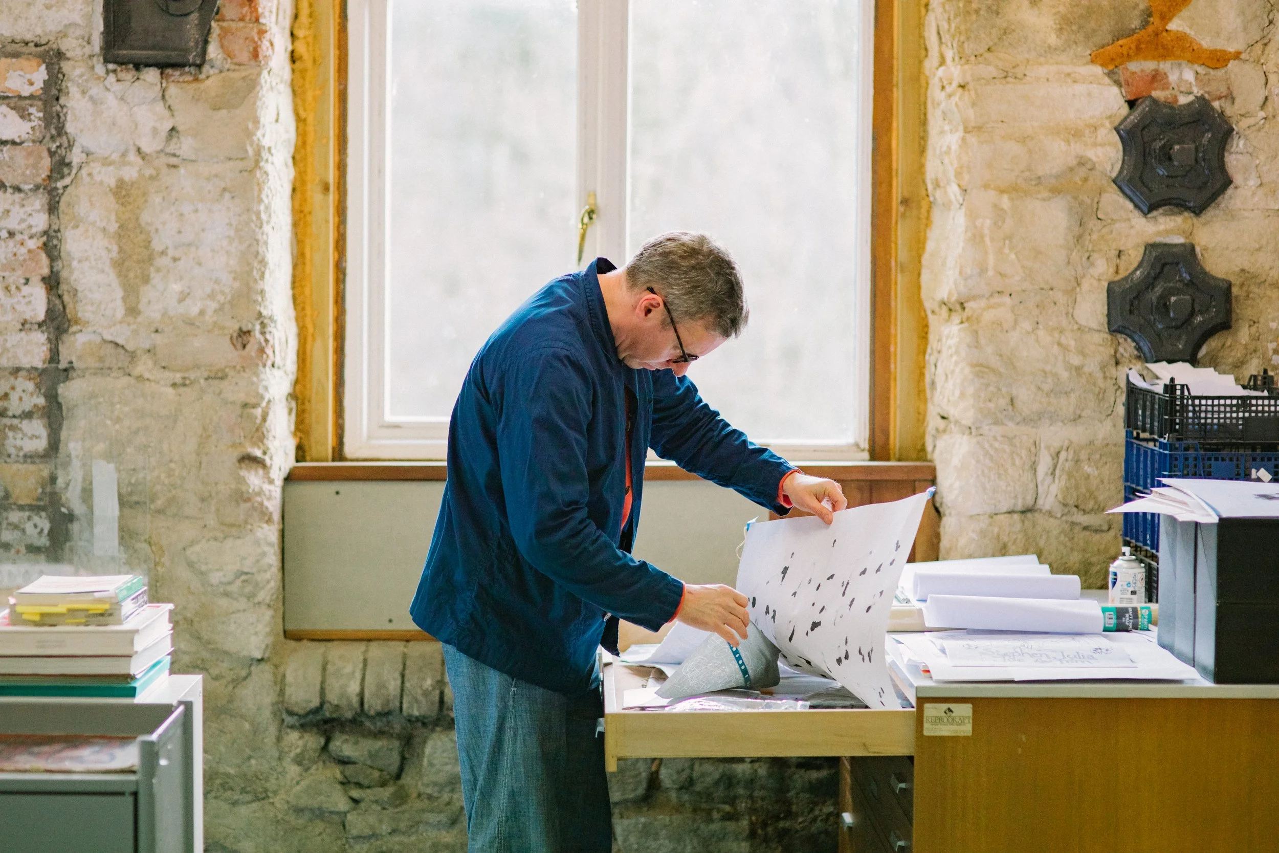 A man wearing glasses and a blue jacket stands at a table looking at and organizing papers or posters in a room with stone walls and a window.