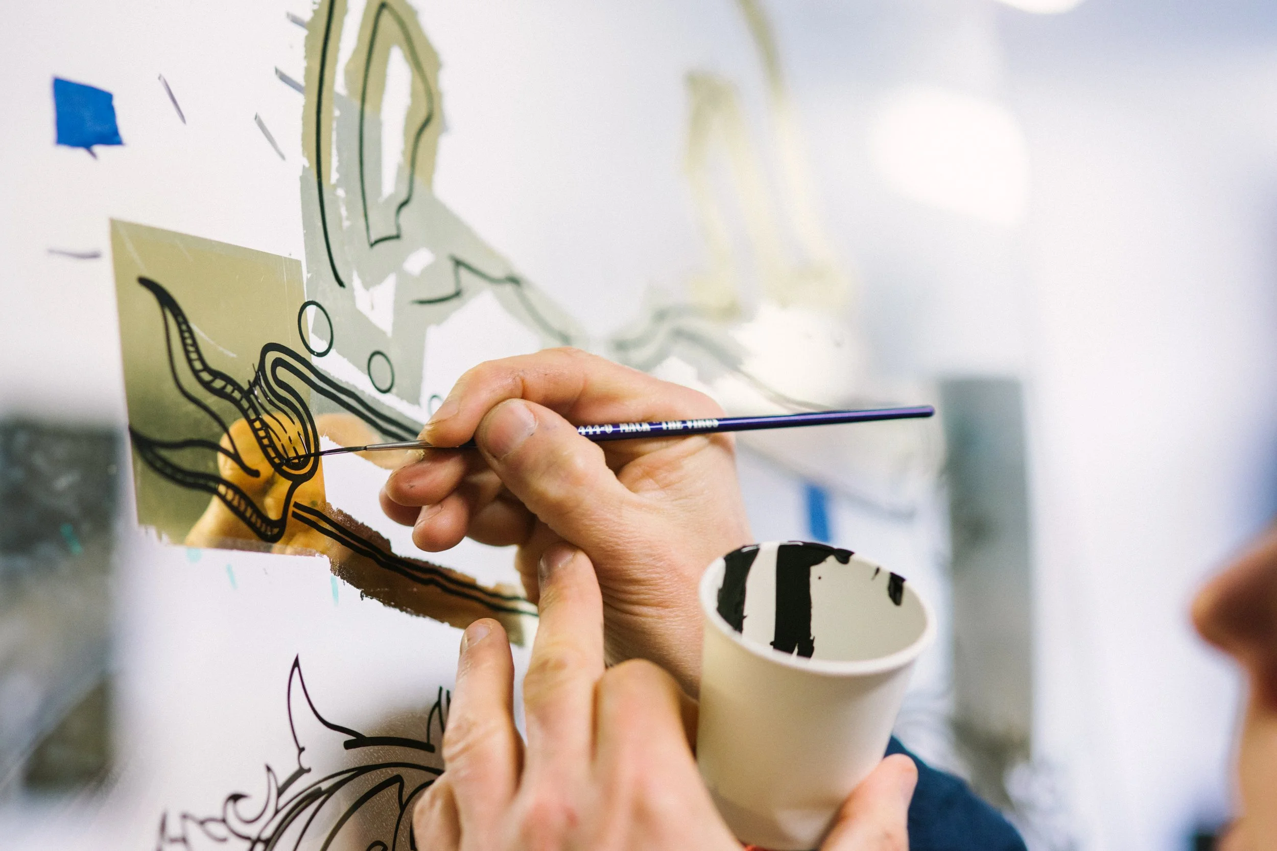 A person uses a fine-tipped brush to paint a detailed black outline of a map on a transparent surface, with a paper cup holding the paint nearby.