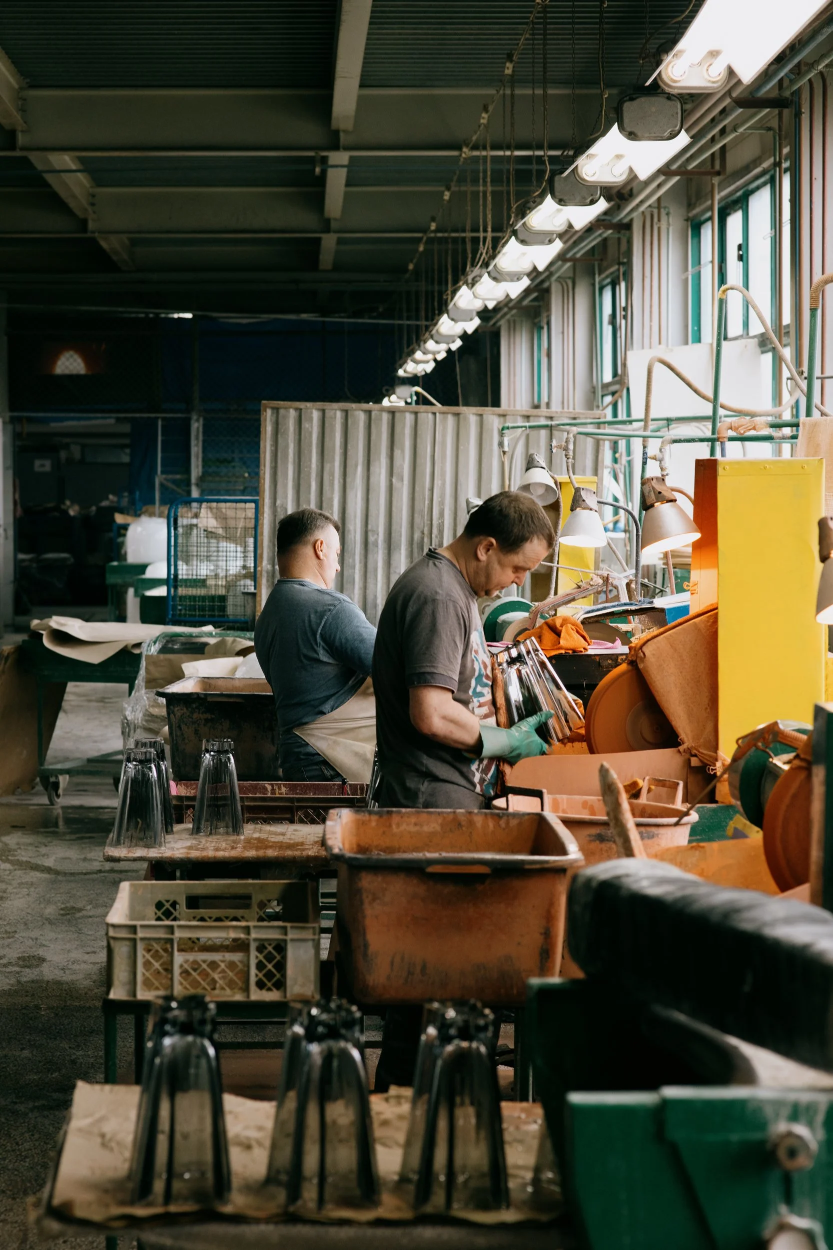 Two workers in a factory or workshop working with reindeer antlers and glassware, overcast lighting, inside a warehouse with high ceilings and large windows.