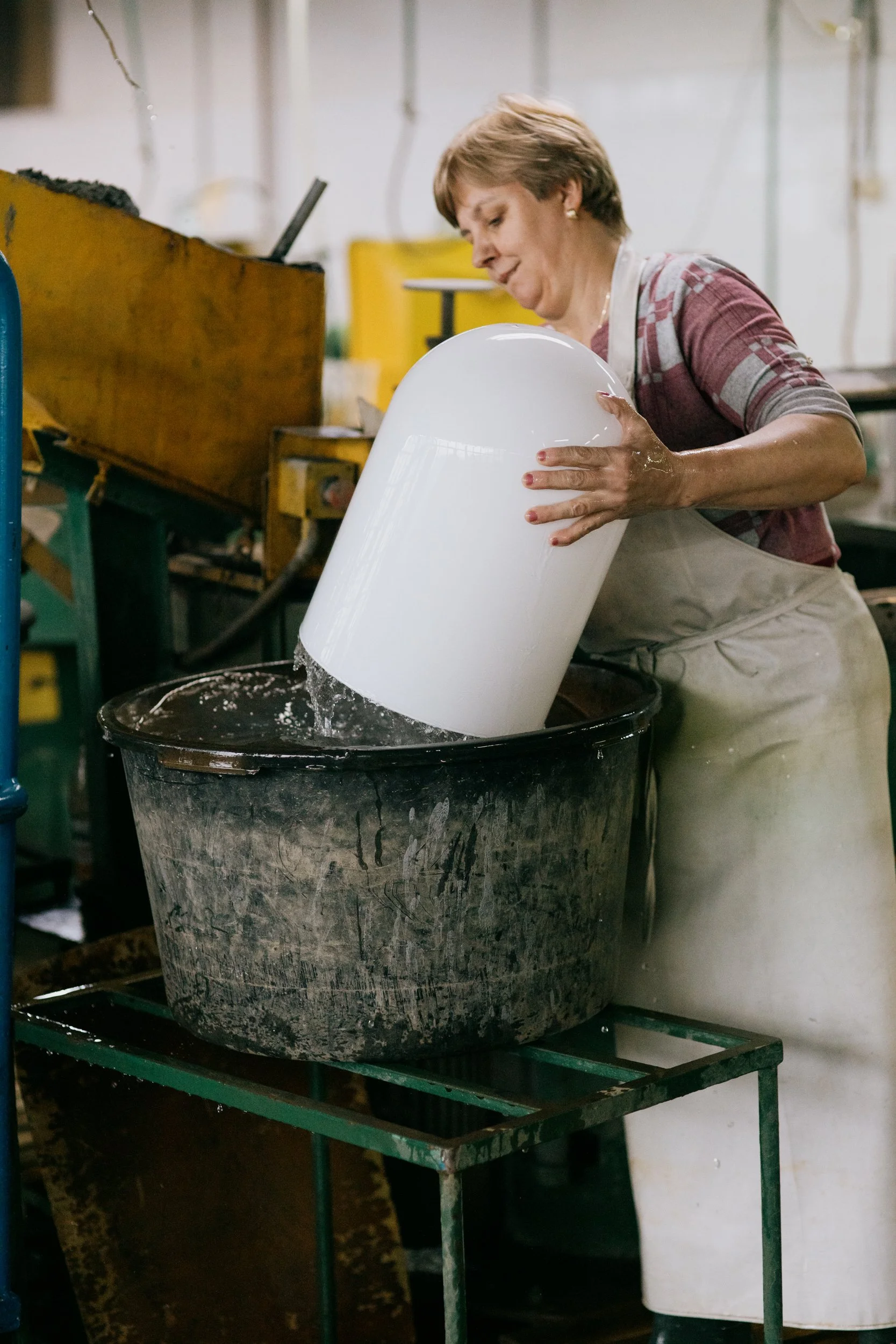A woman in apron pressing a large white object, possibly porcelain or ceramic, into a barrel or container with water in an industrial or workshop setting.