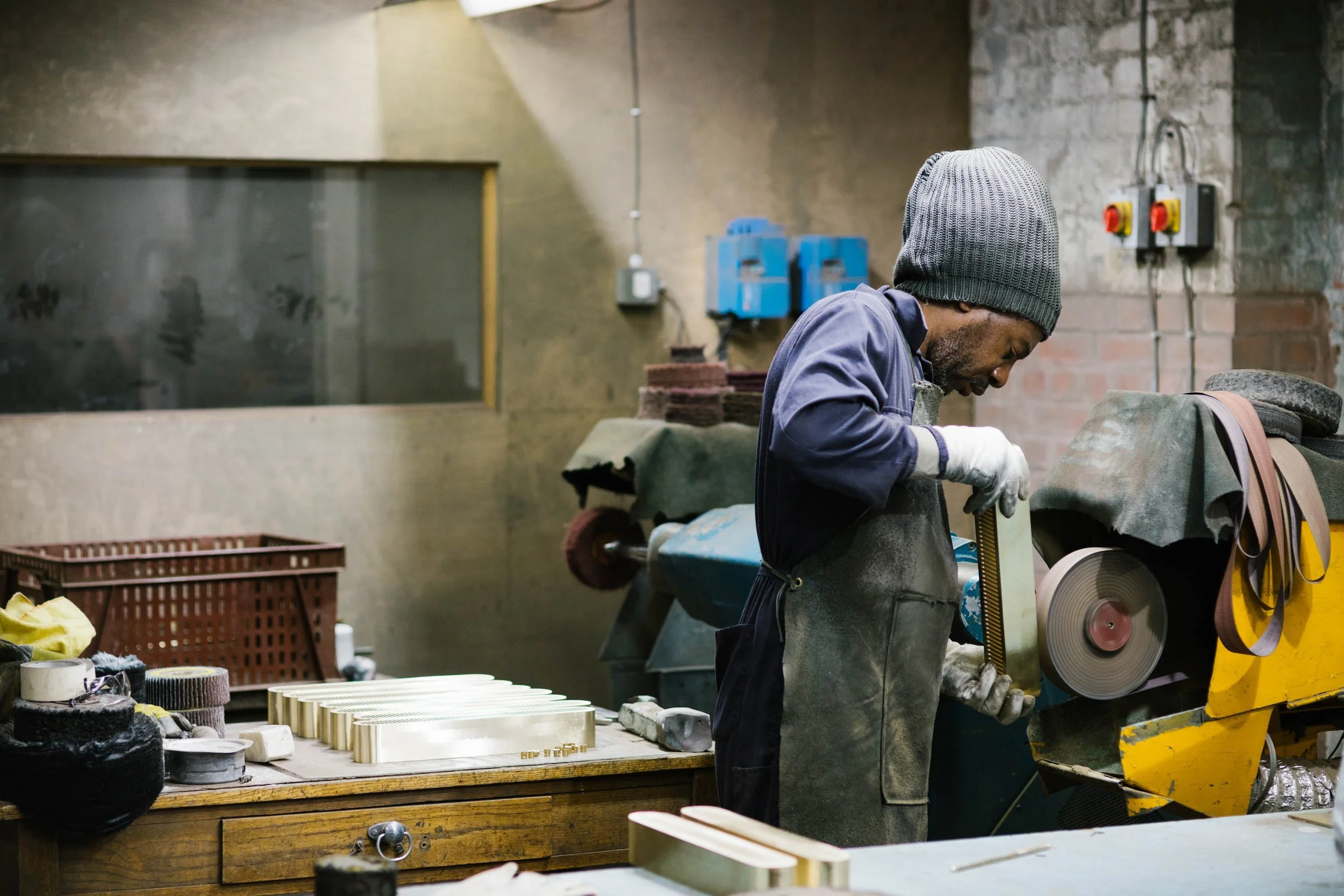 A craftsman wearing a gray beanie and gloves works with tools in an industrial workshop with brick and concrete walls, surrounded by wood, fabric, and machinery.