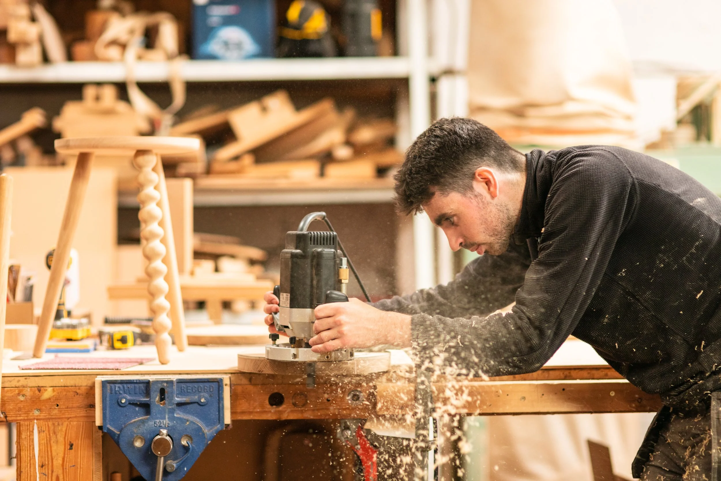 A person working in a woodworking shop, using a piece of machinery on a wooden workbench, surrounded by woodworking tools and wood pieces.