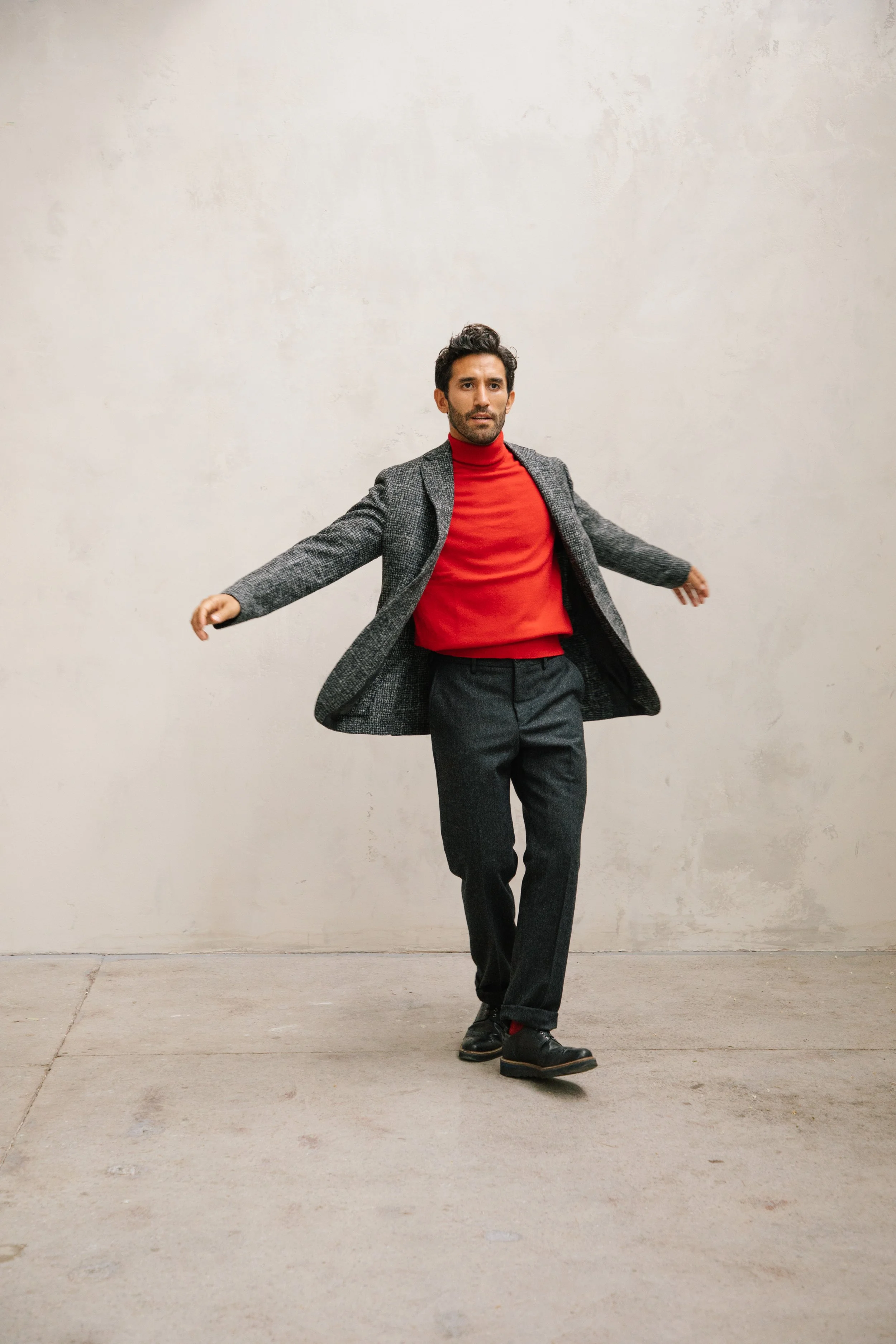 A man walking with arms outstretched, wearing a gray blazer, red turtleneck, dark gray pants, and black shoes, in front of a plain off-white wall.