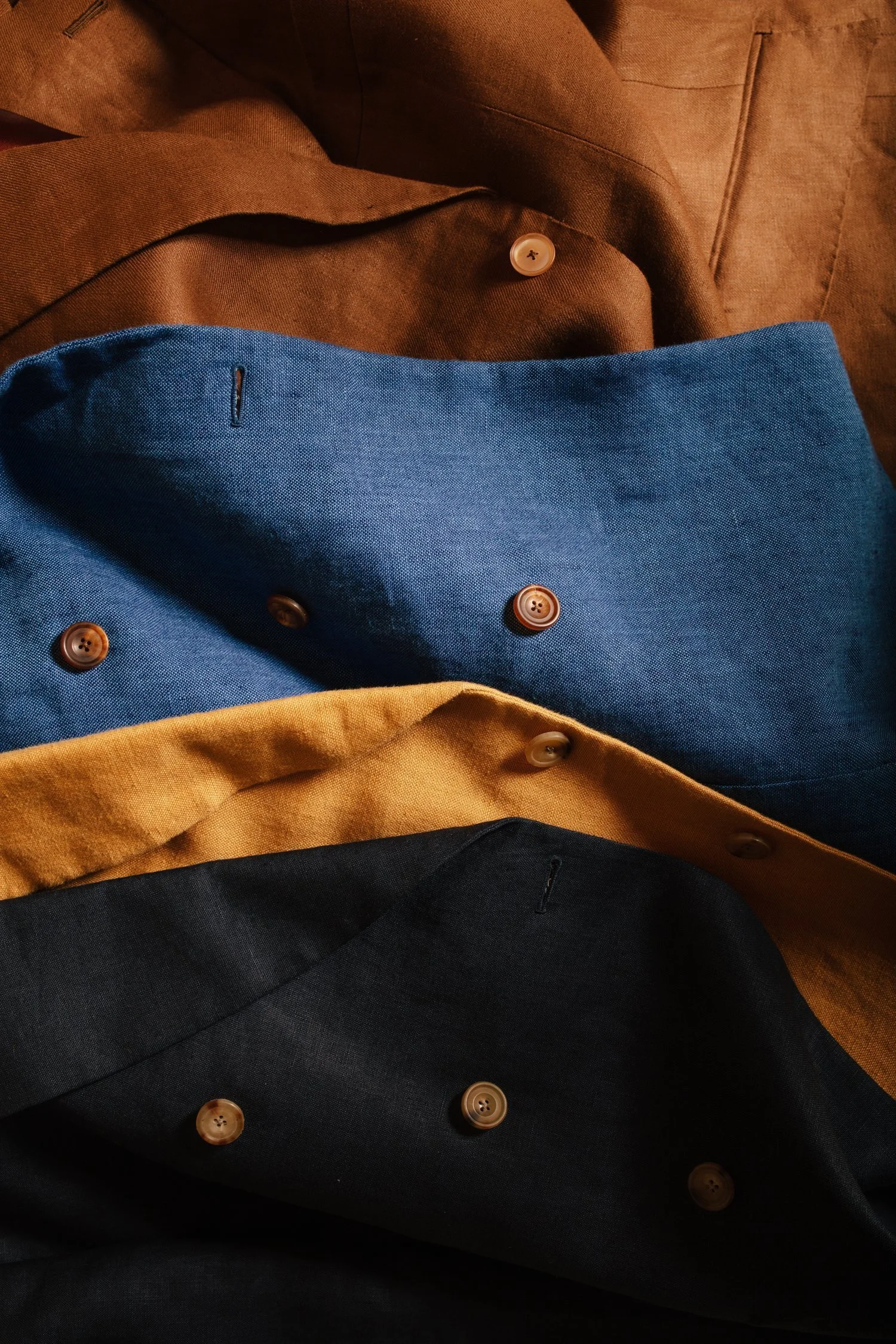 Close-up of five colorful shirts layered on top of each other with visible buttons, including brown, blue, yellow, and dark blue fabrics.