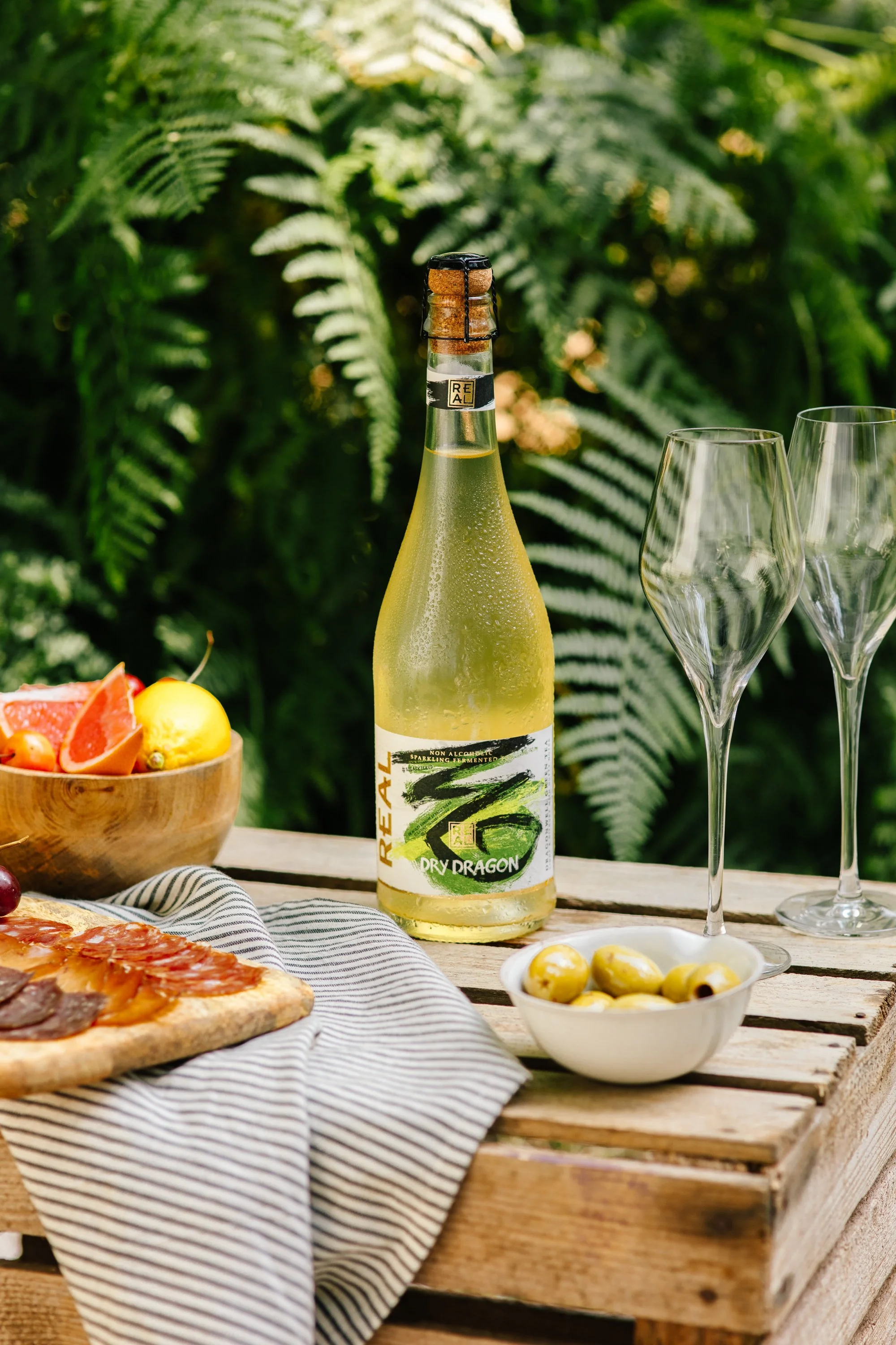 A picnic table with a bottle of dry ginger beer, a bowl of green olives, a plate with sliced cured meats, and a pizza on a striped cloth, set outdoors with lush green foliage in the background.