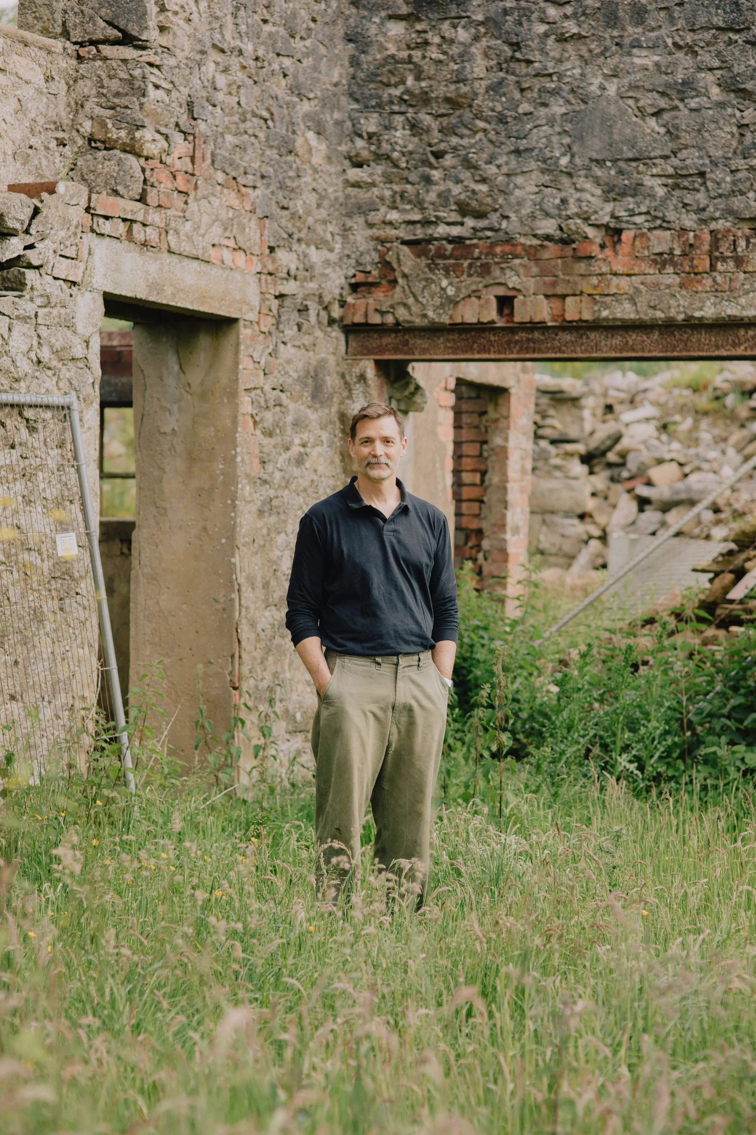 A middle-aged man with short brown hair and a beard stands outdoors in front of a weathered, abandoned building with brick and stone walls and a rusted metal roof. He is wearing a black long sleeve shirt and khaki pants, with his hands in his pockets