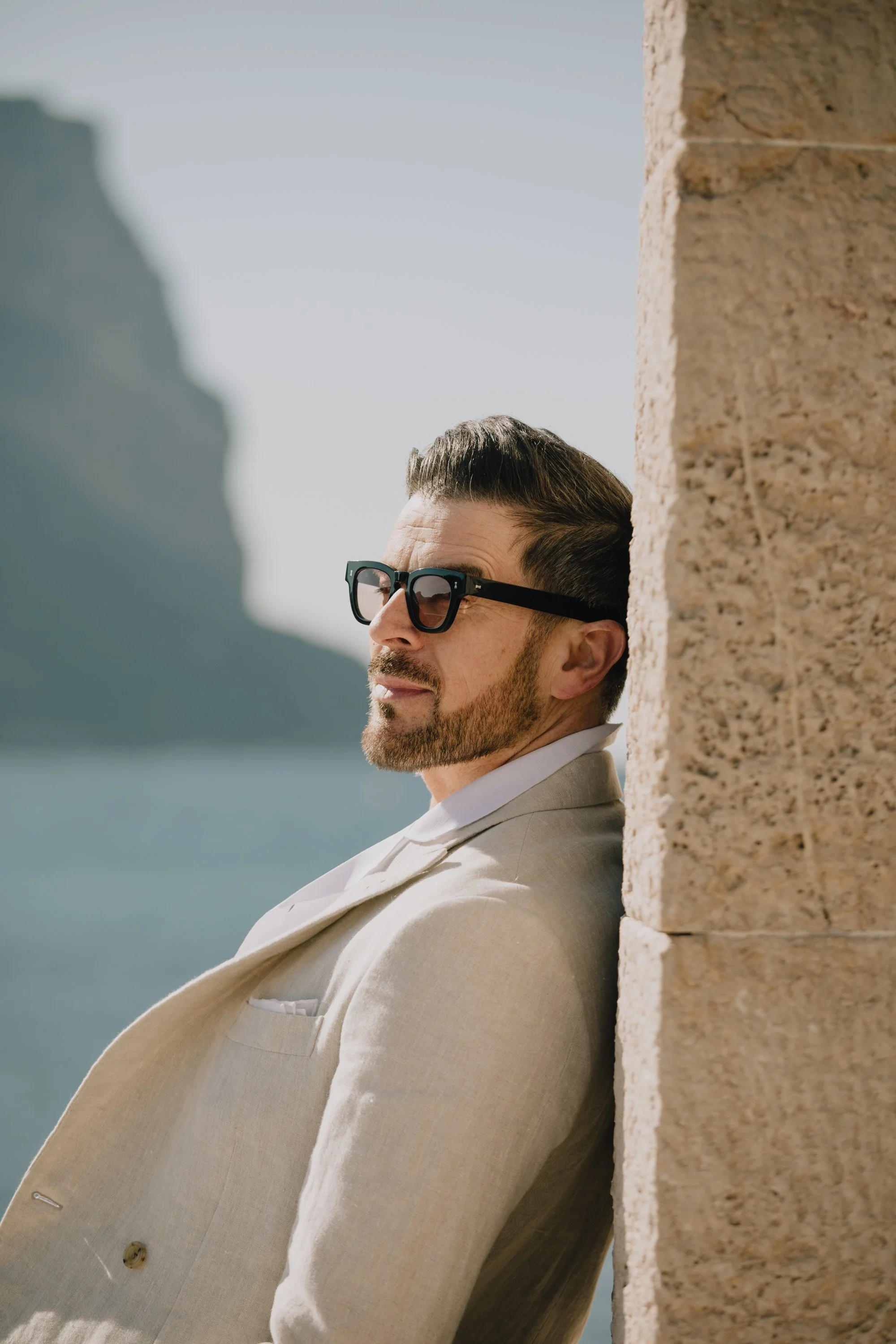 A man in a beige suit and sunglasses leaning against a stone wall, outdoors with mountains and water in the background.