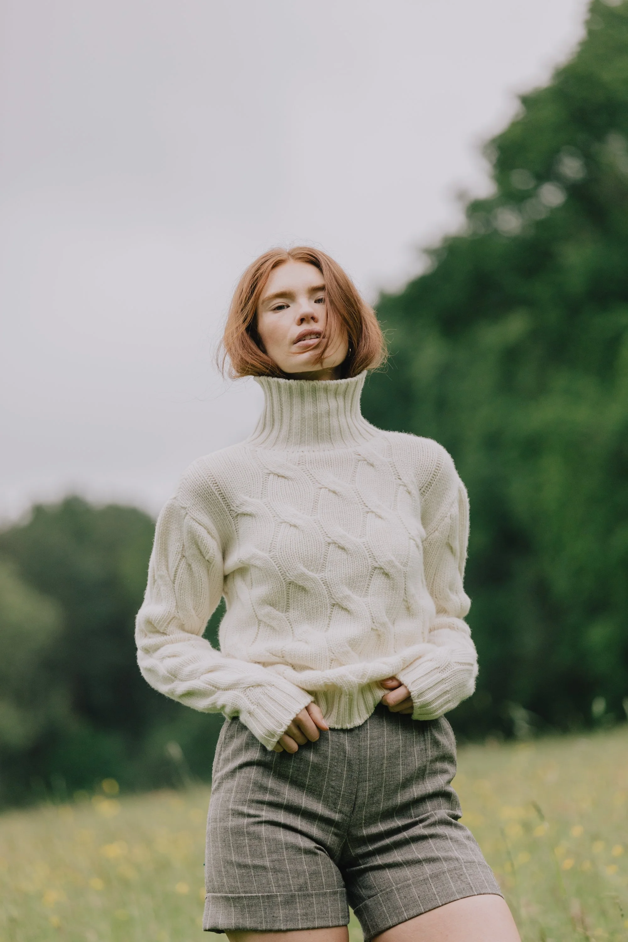 A woman with red hair wearing a cream-colored turtleneck sweater and gray plaid shorts standing outdoors in a grassy area with trees in the background.