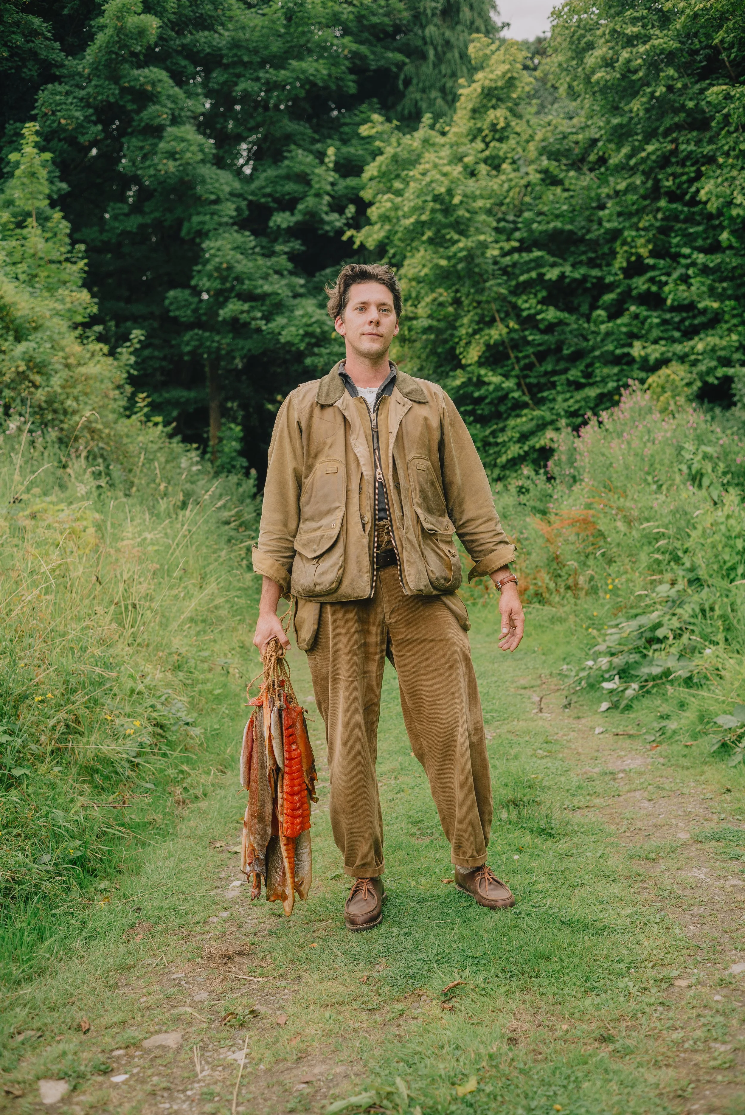 A man in outdoor clothing standing on a grassy trail in a lush green forest. He holds a fishing net with fish in one hand and looks directly at the camera.