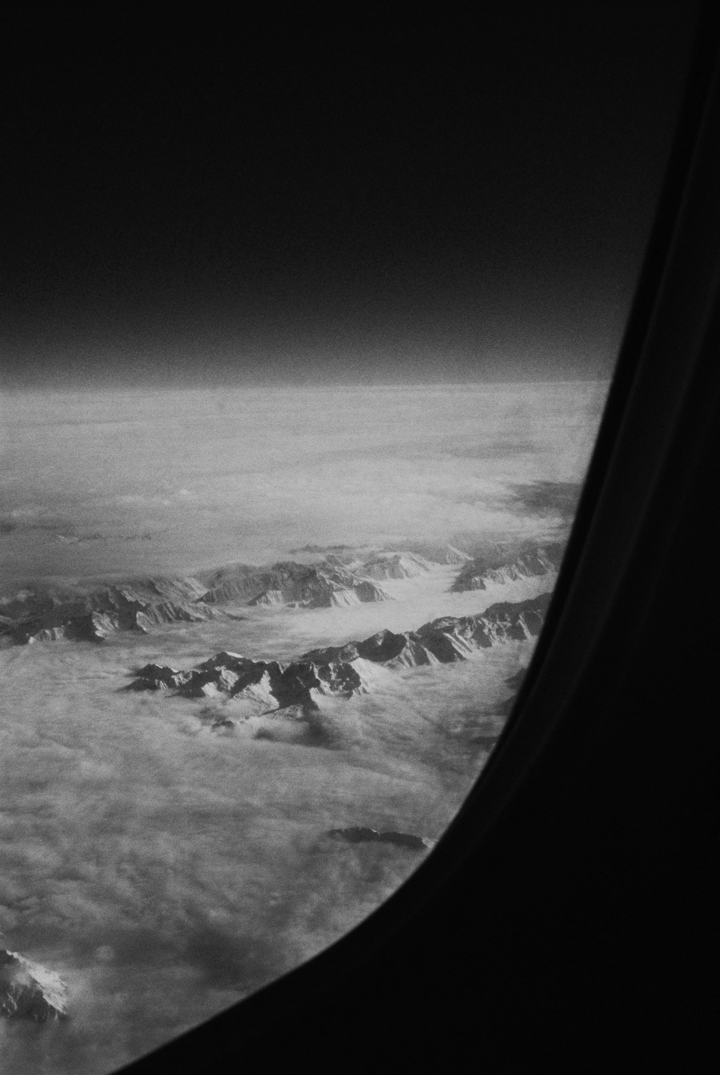 Black and white aerial view from an airplane window showing mountain peaks and glacier fields below, with a dark sky above.