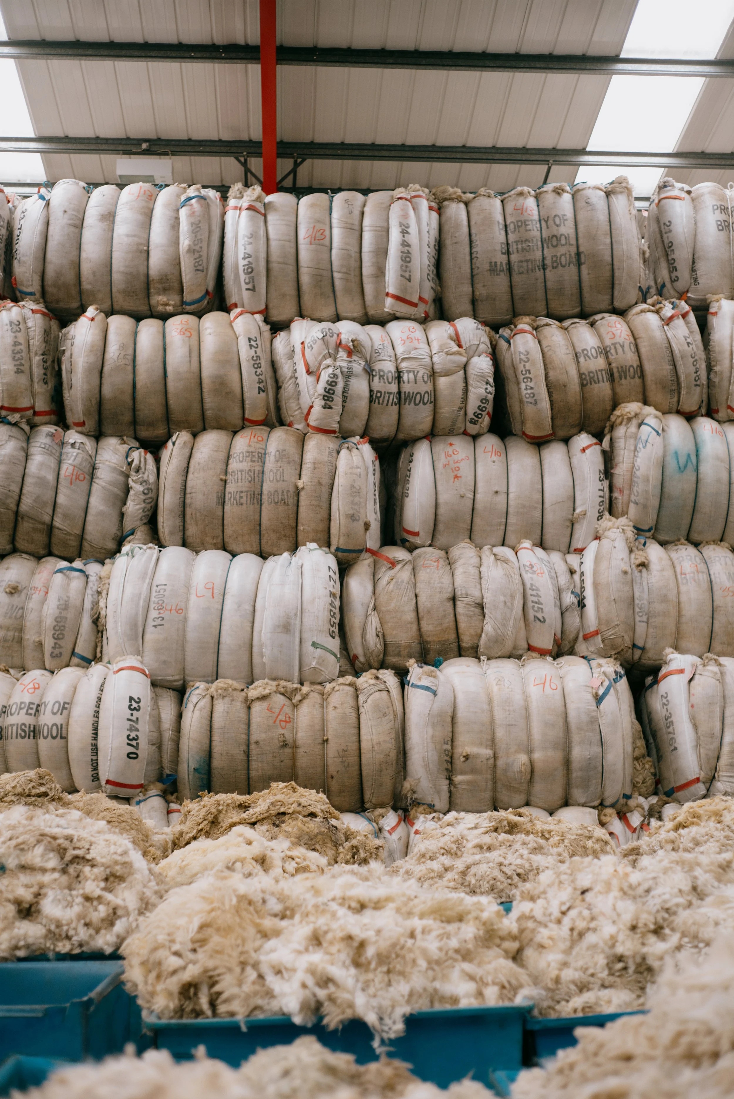 Stacks of white bags filled with wool stored on shelves in a warehouse, with loose wool fibers on the floor below.