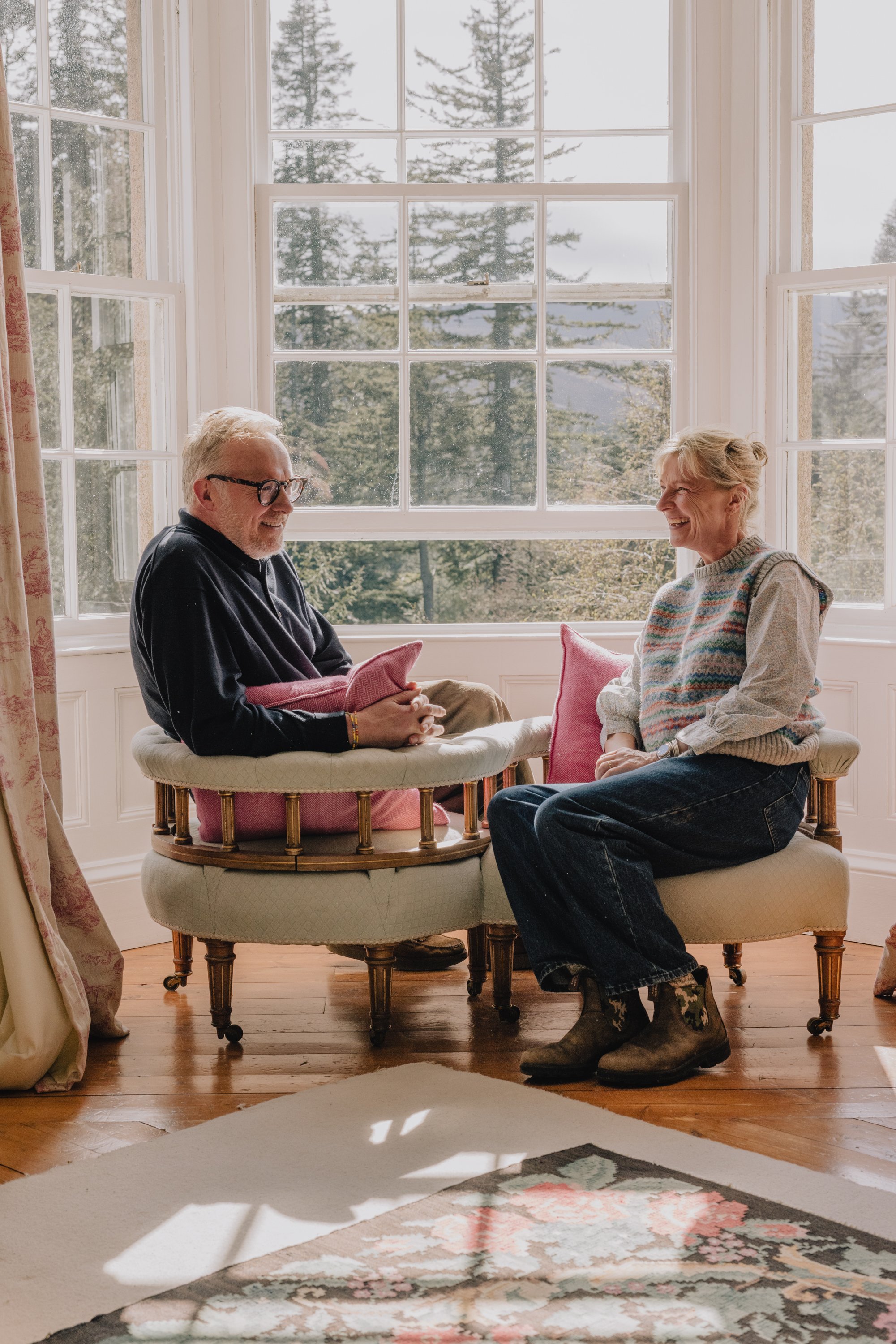 Two people, an older man with glasses and a beard, and an older woman with blonde hair, are sitting in a sunlit room with large windows behind them. They are smiling and facing each other, engaged in conversation, with colorful cushions around them a