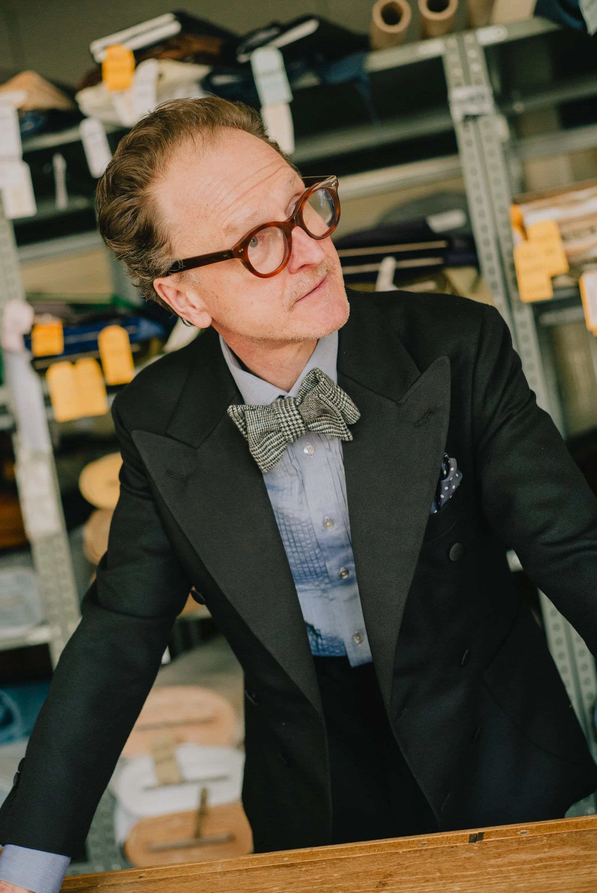 A man wearing a black suit and bow tie with round glasses, working or studying at a wooden table in a workshop or office setting with shelves and supplies in the background.