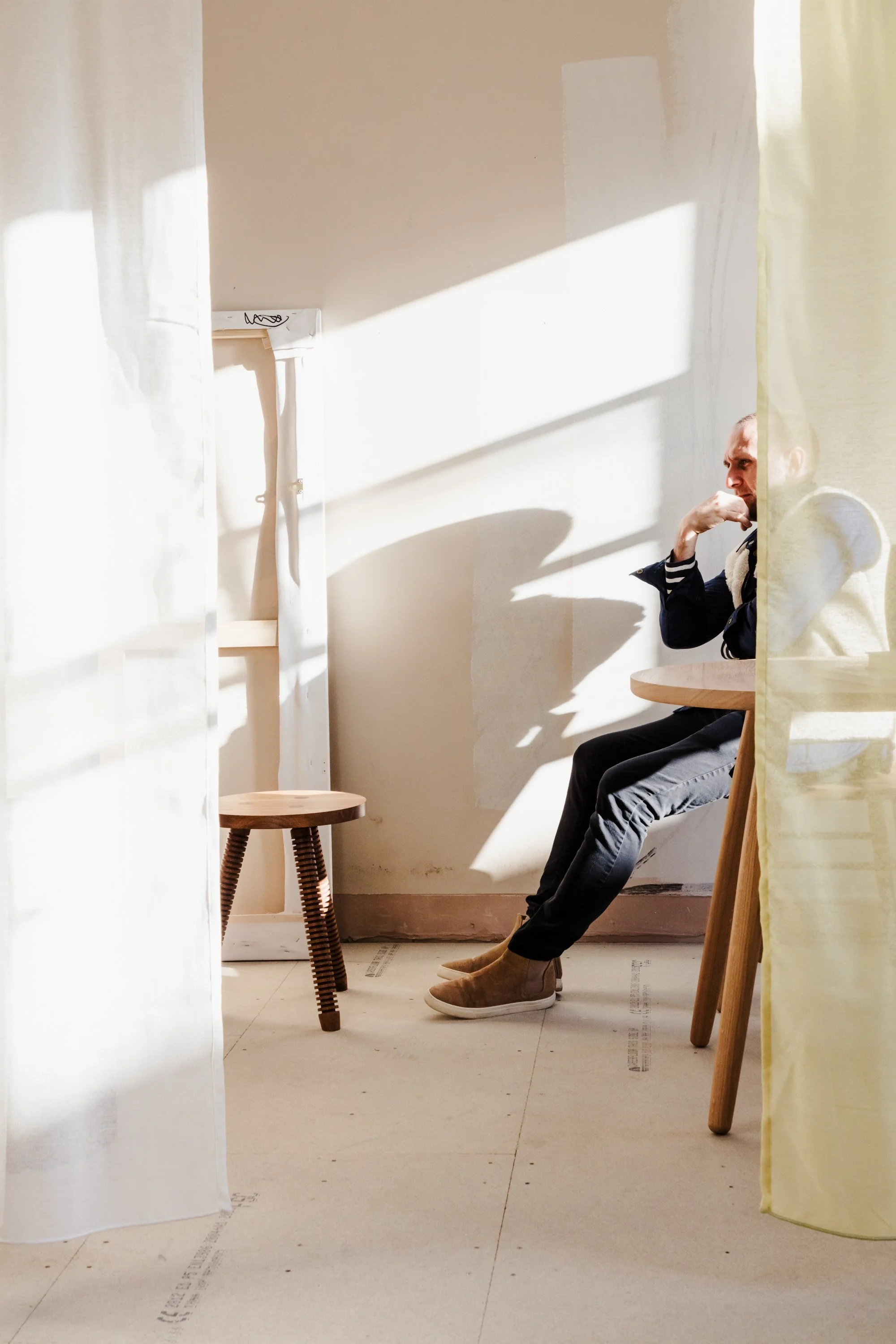 A man sitting on a wooden chair, partially obscured by yellow curtains, is engaged in a thoughtful pose with his chin resting on his hand. He is wearing a black jacket, gray pants, and beige shoes, with sunlight streaming through a window creating sh