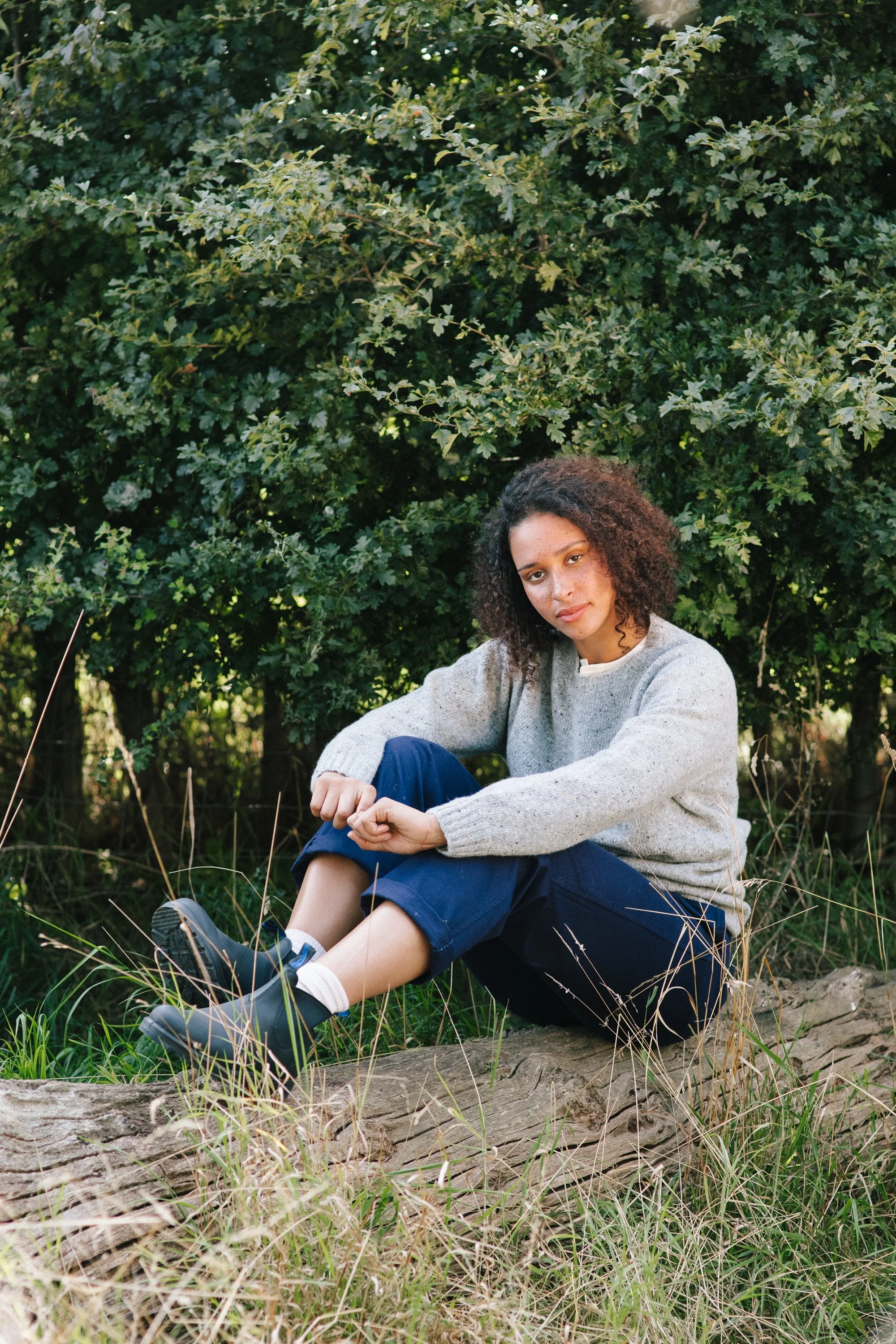 A young woman with curly hair and freckles posing outdoors on a log in a natural setting with green foliage and grass.