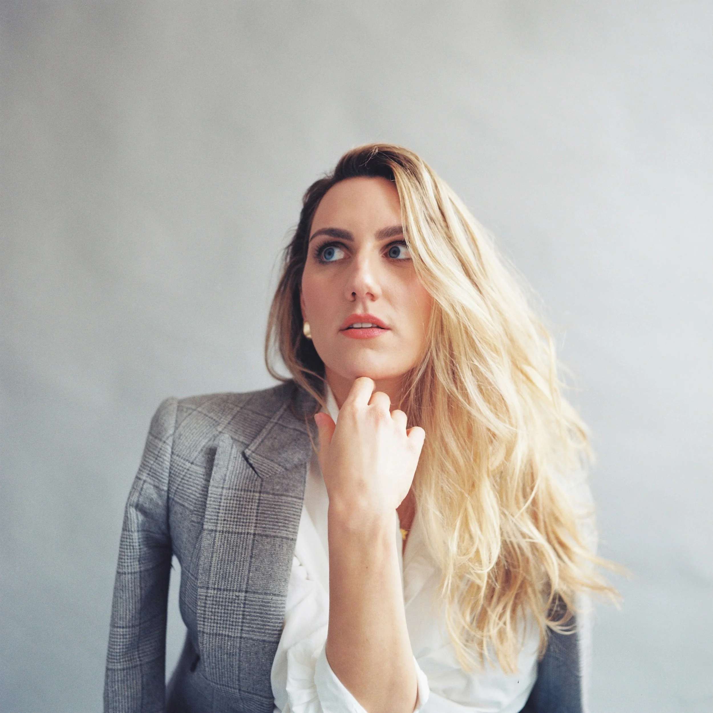 A woman with long, wavy blonde hair and blue eyes wearing a white shirt and a gray plaid blazer, with her hand touching her chin, looking slightly upward.