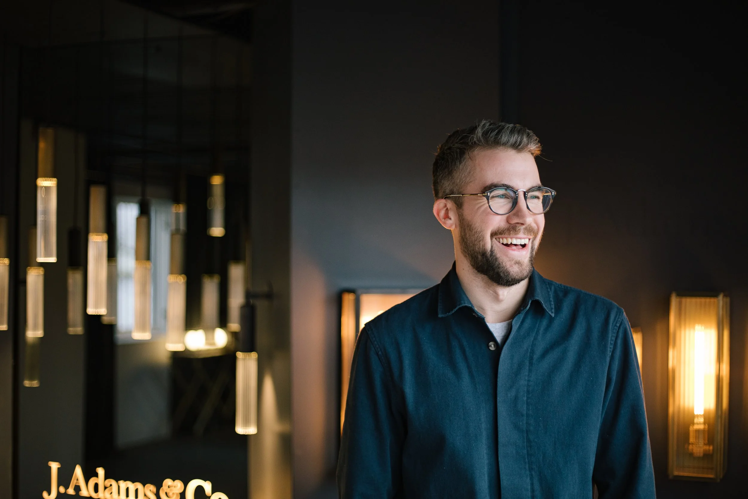 A smiling man wearing glasses and a dark blue shirt, standing in a modern interior with decorative lighting and a sign that says "J. Adams & Co."