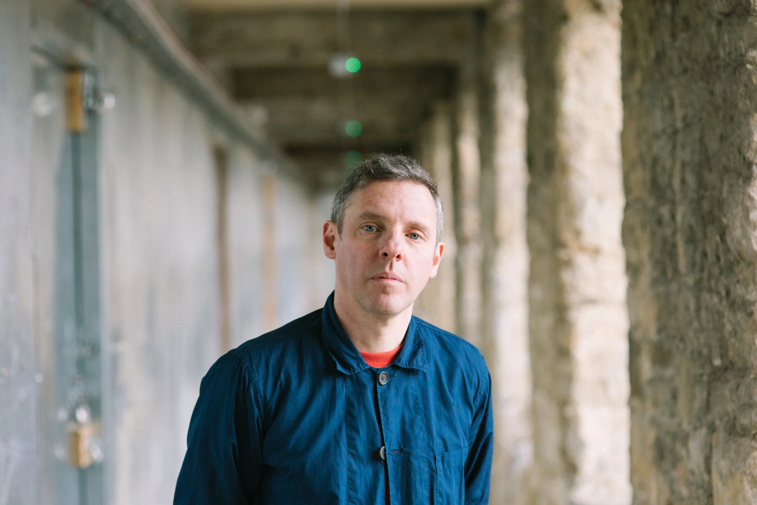 A man with short hair and a serious expression, wearing a dark blue button-up shirt, standing in front of a textured stone wall with an industrial background.