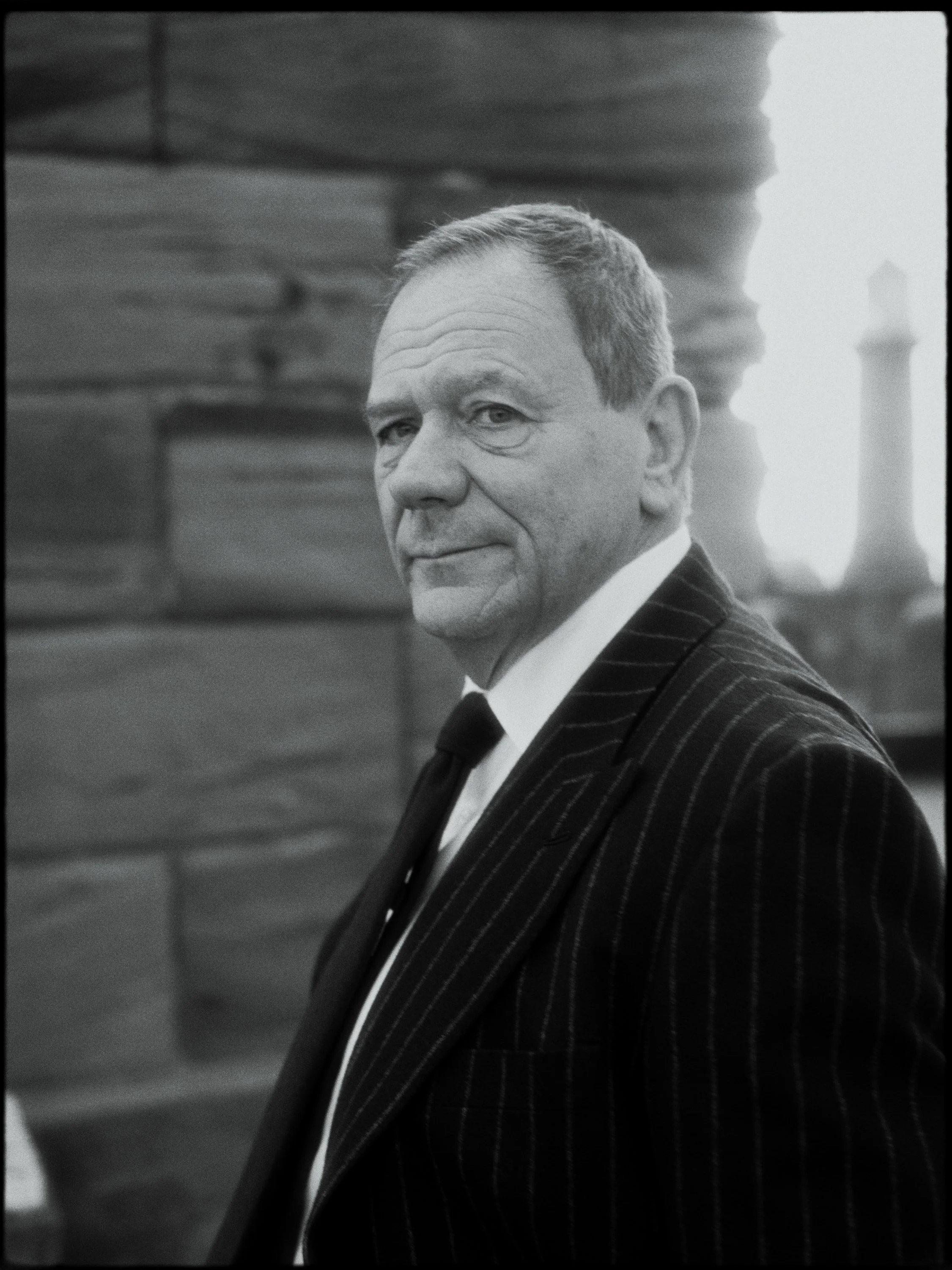 A black-and-white photo of a man in a pinstripe suit looking over his shoulder with a slight smile, outdoors near a body of water with a large lighthouse in the background.