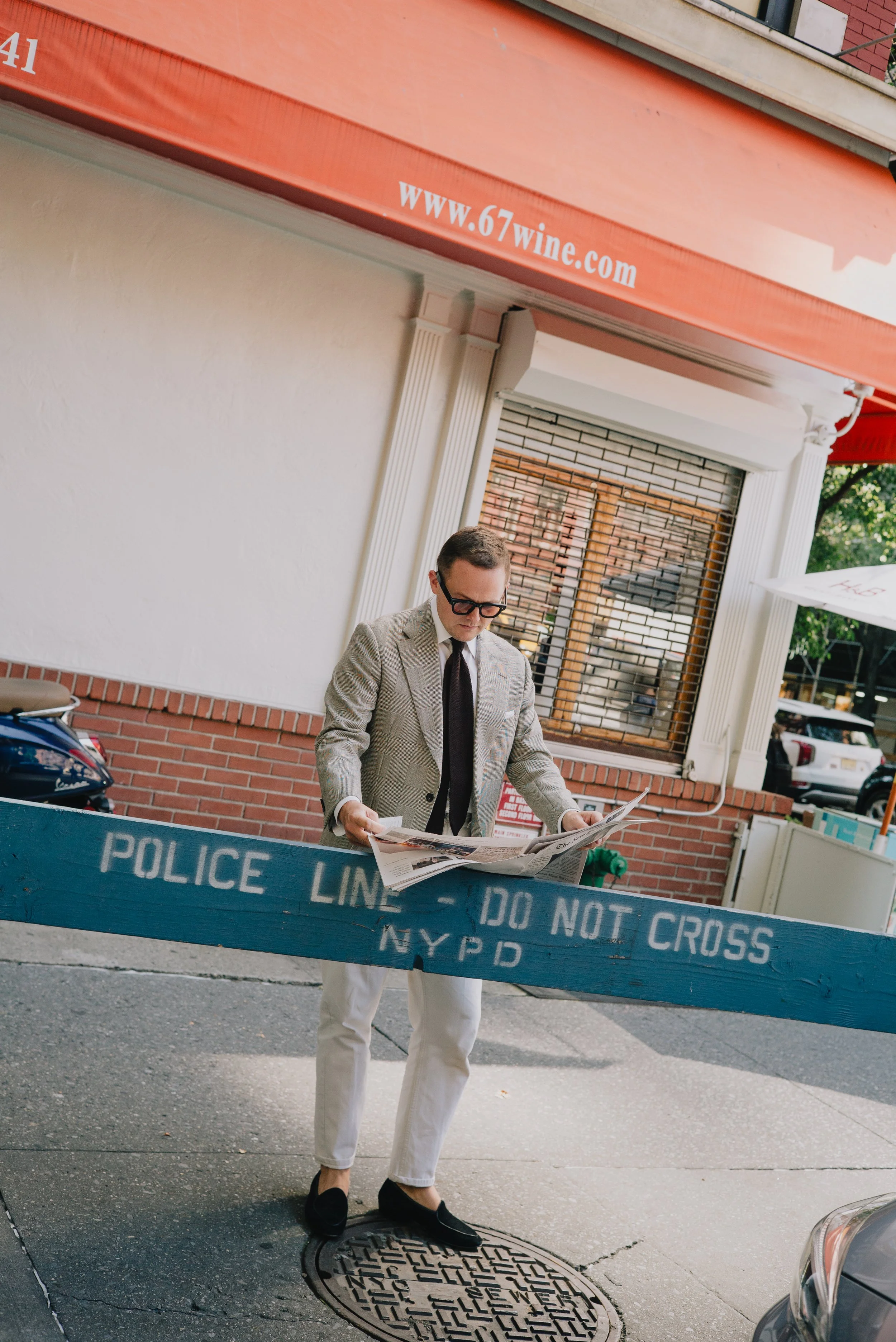 A man in a beige checkered blazer, black tie, and white pants is standing on a city sidewalk, reading a newspaper. He has dark glasses and black loafers. Behind him, there is a blue police barricade with white text that reads 'POLICE LINE DO NOT CROS