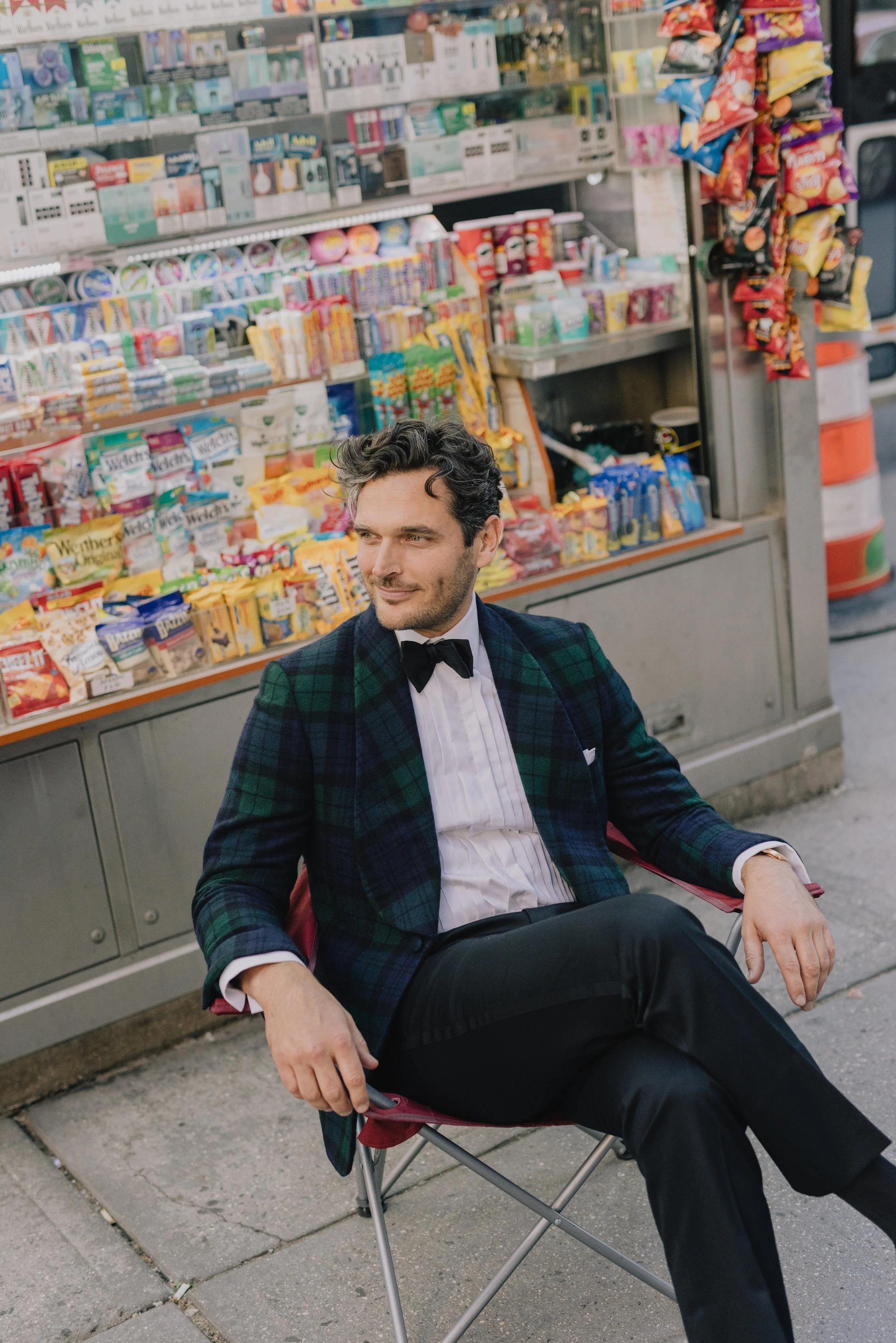 A man dressed in a checkered blazer, white shirt, and black bow tie sitting in a folding chair outside a convenience store. The background shows shelves stocked with snacks and candies.