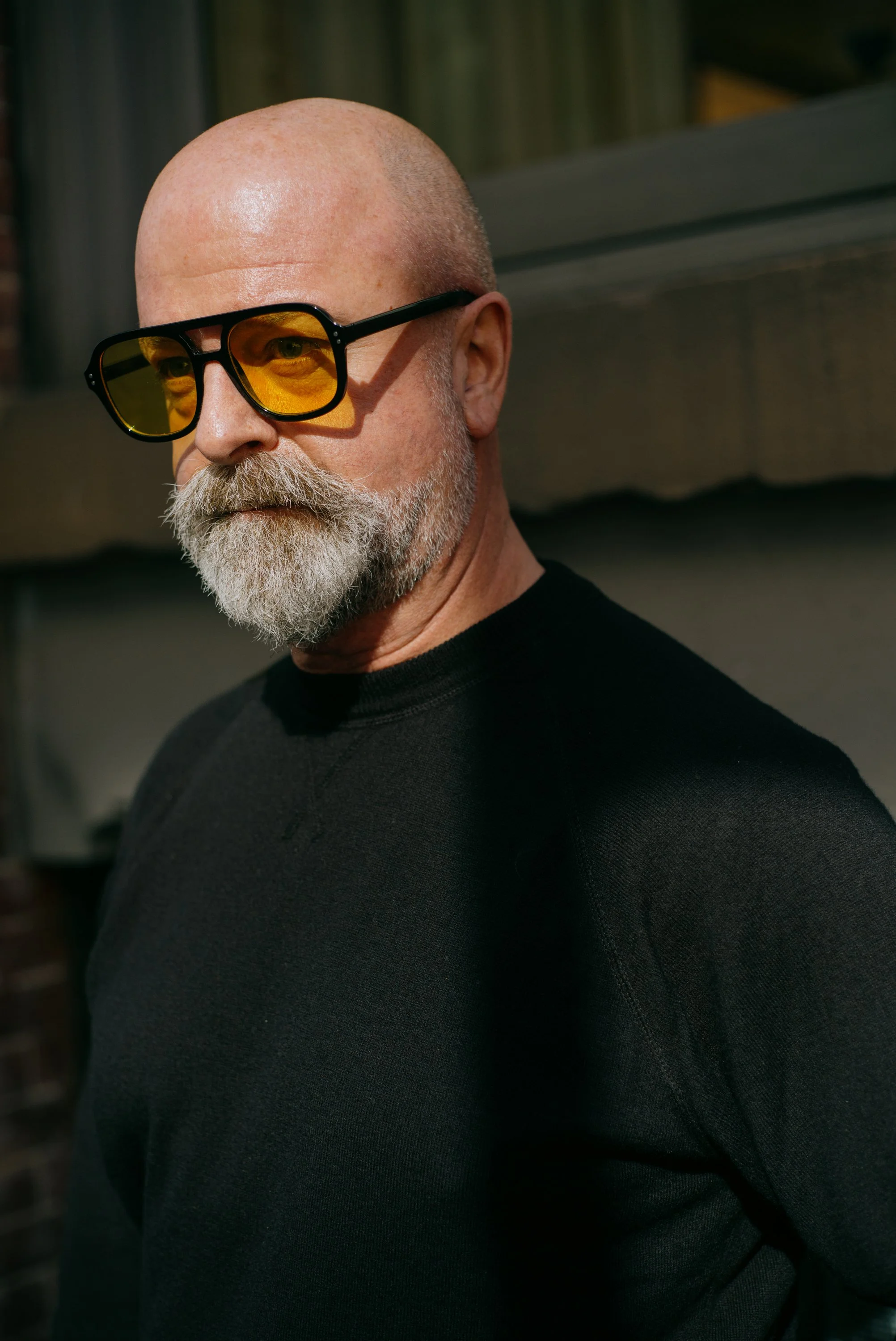 A bald man with a white beard wearing yellow-tinted sunglasses and a black shirt.