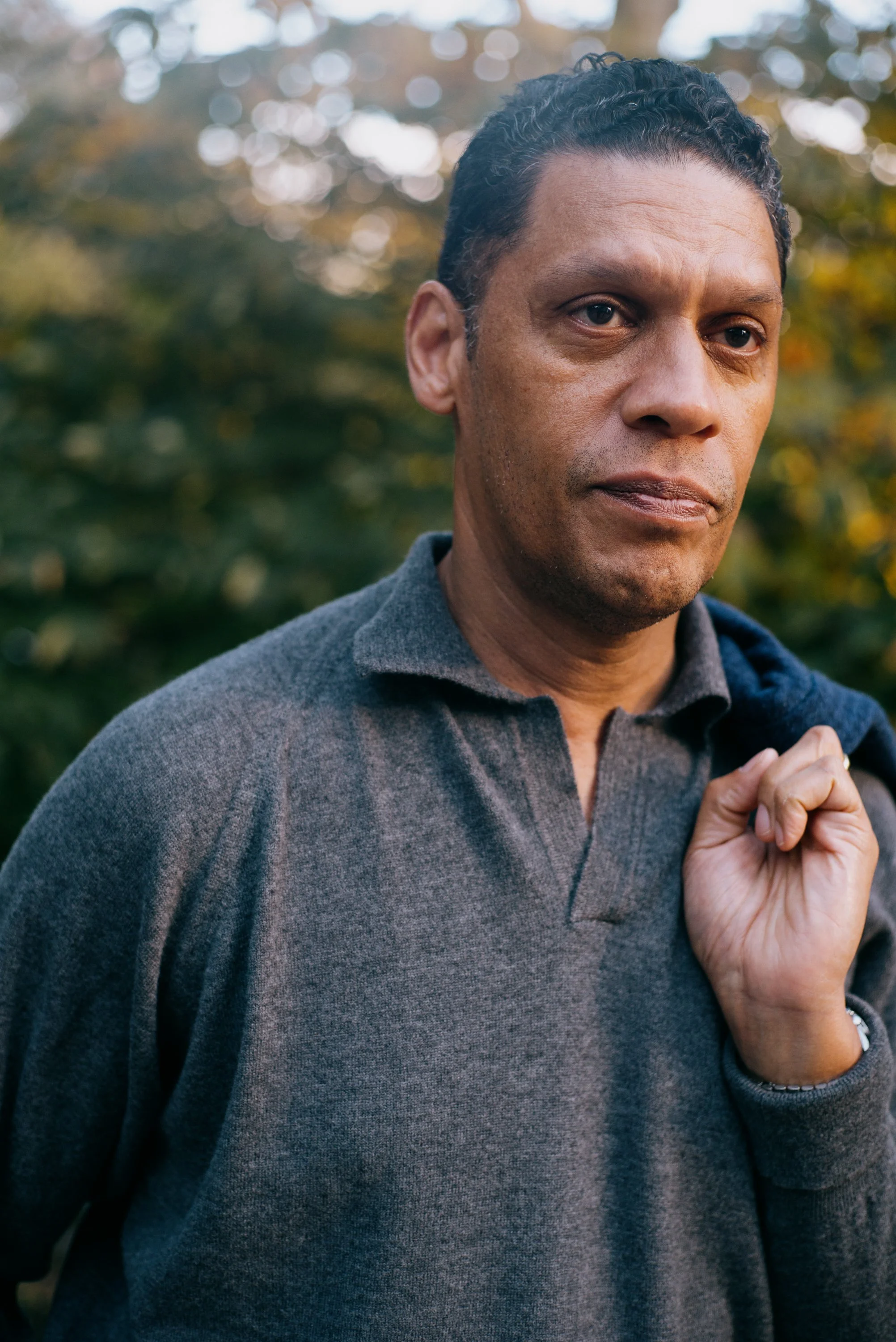 Close-up of a middle-aged man with short, curly dark hair wearing a gray sweater, standing outdoors with blurred autumn trees in the background.