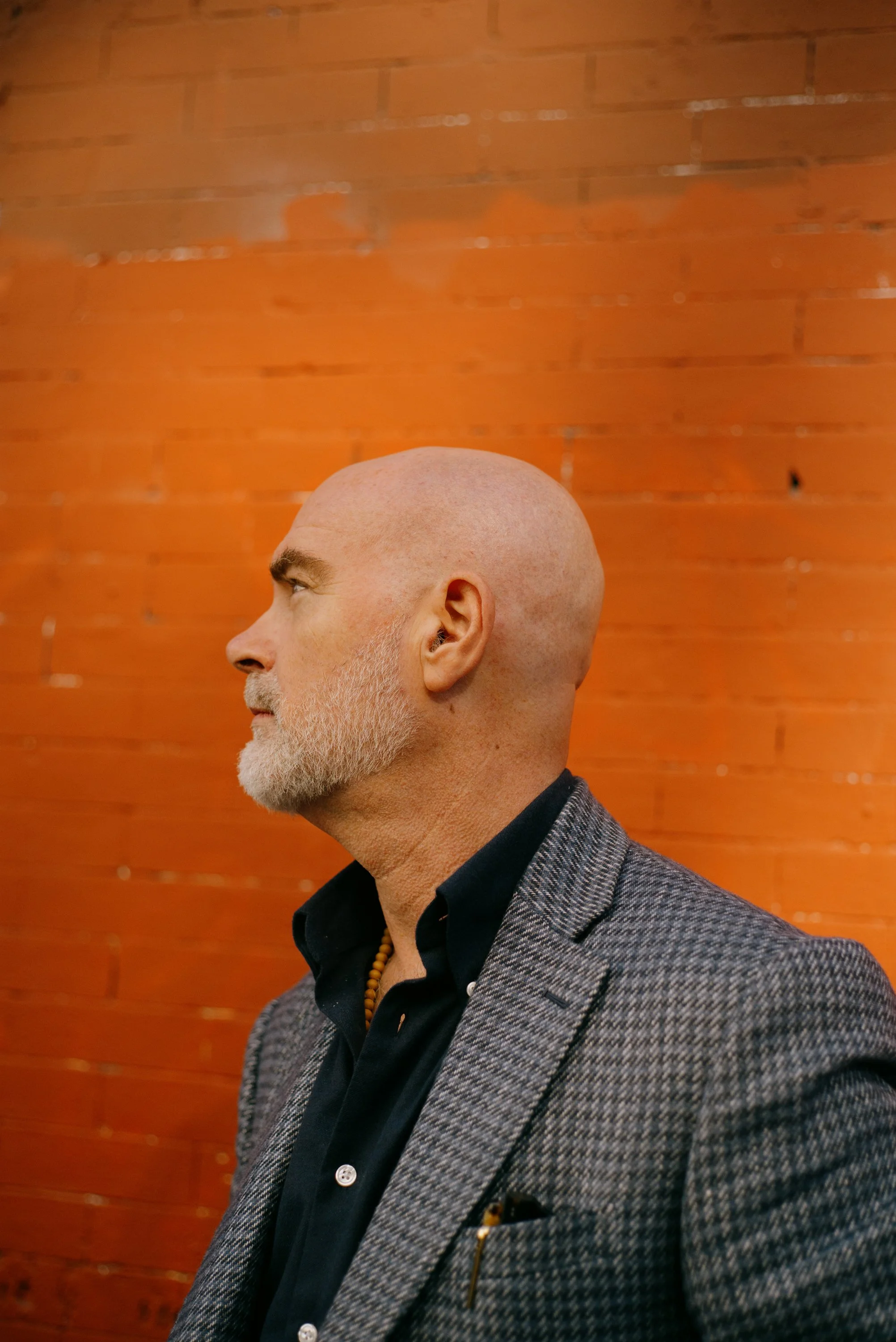 Profile of an older man with a bald head and a gray beard, wearing a gray checked jacket and a black shirt, standing against a wooden wall background.